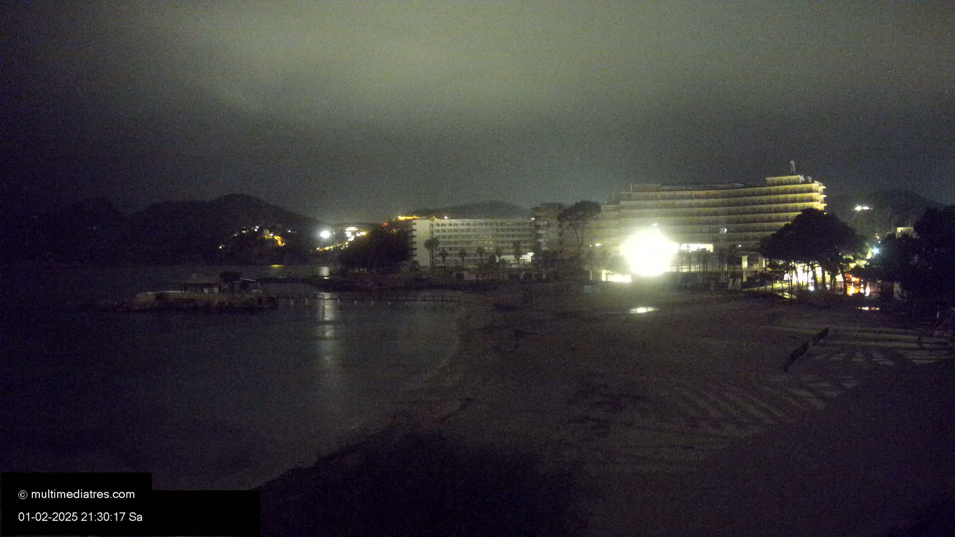 A nighttime view of a beach with a large hotel and other buildings illuminated against a dark, cloudy sky.