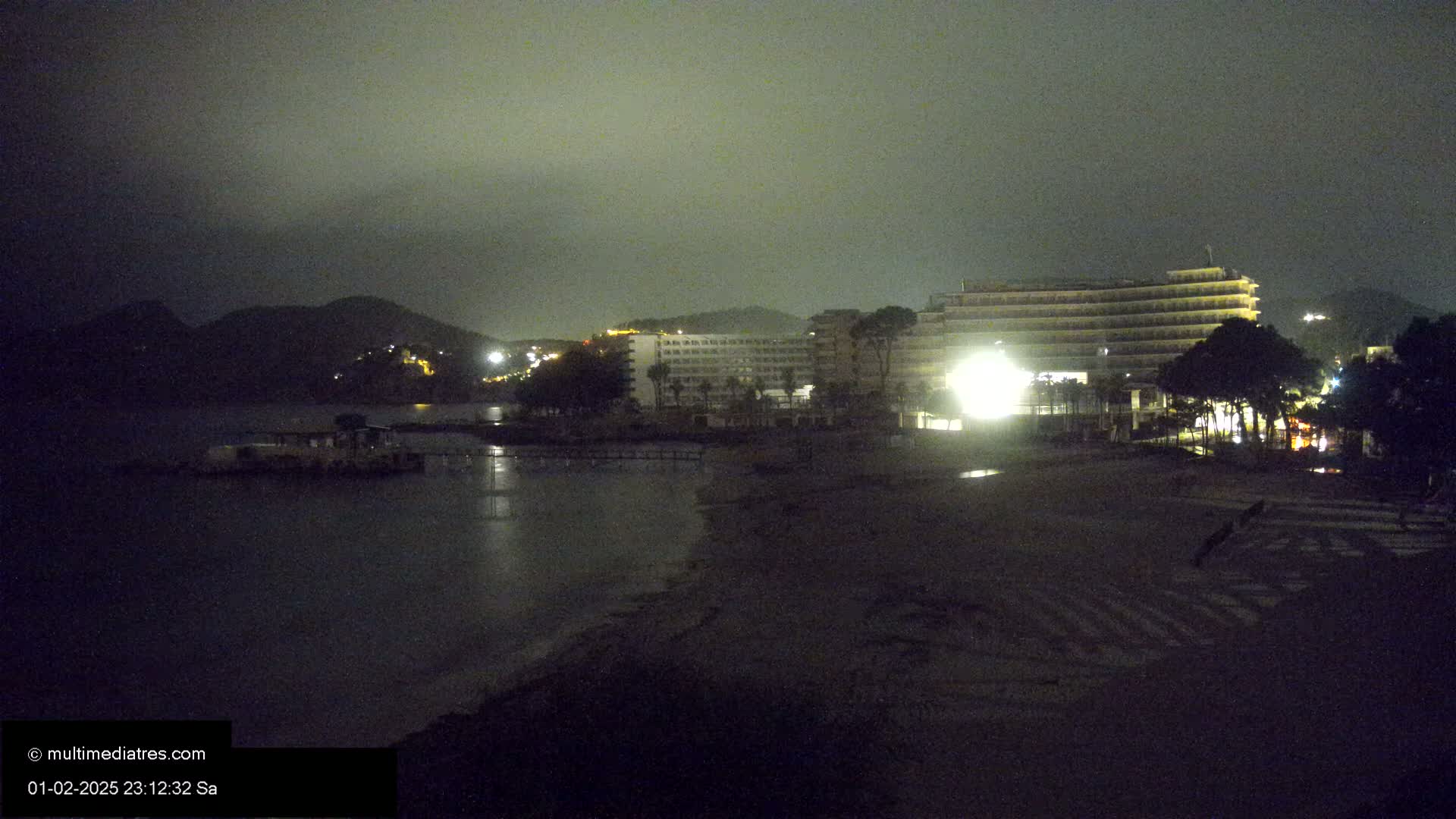 A nighttime view of a beach with a large hotel and hills in the background under a cloudy sky.