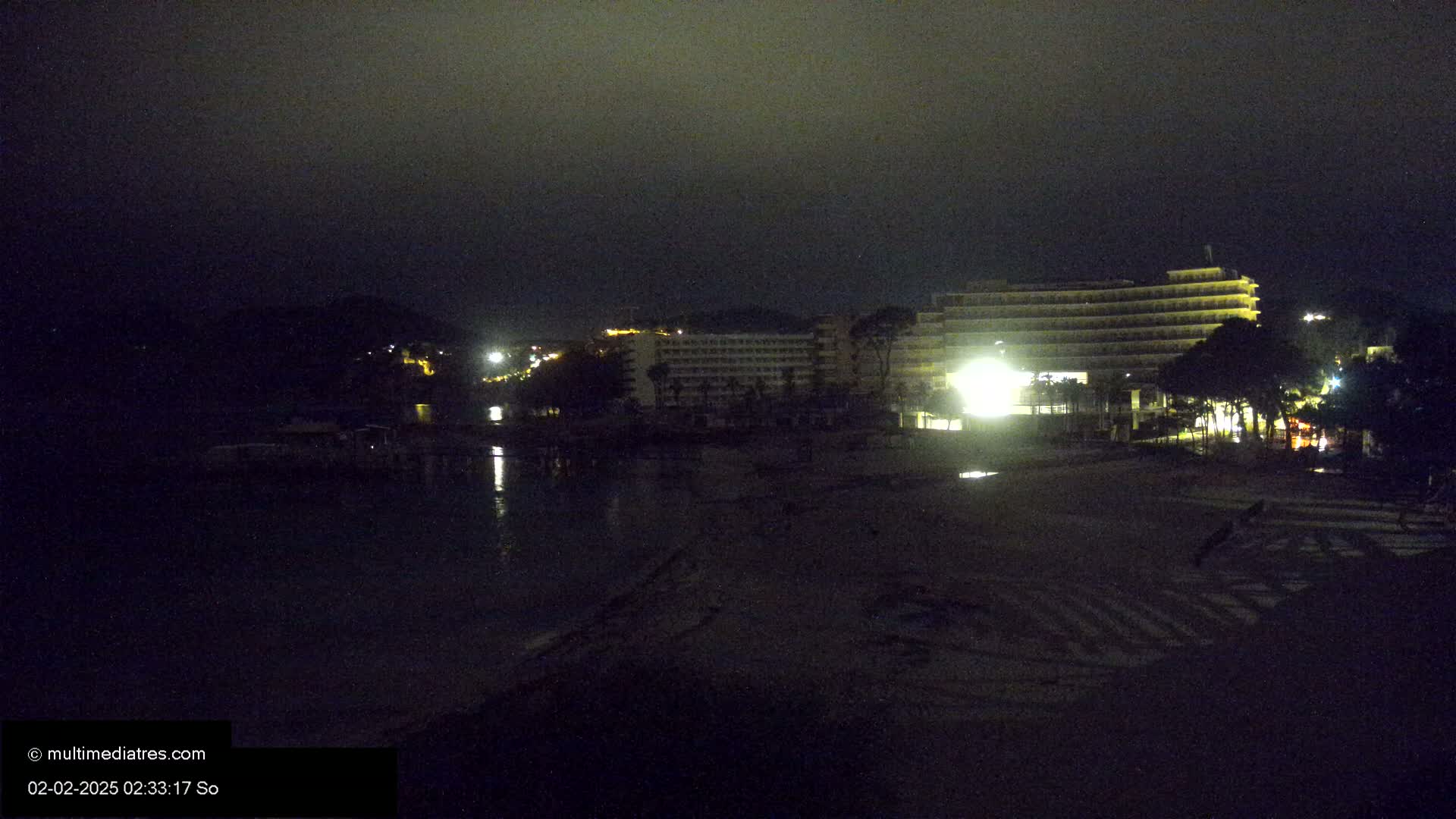A dark, nighttime beach scene shows a multi-story hotel illuminated against a cloudy sky.