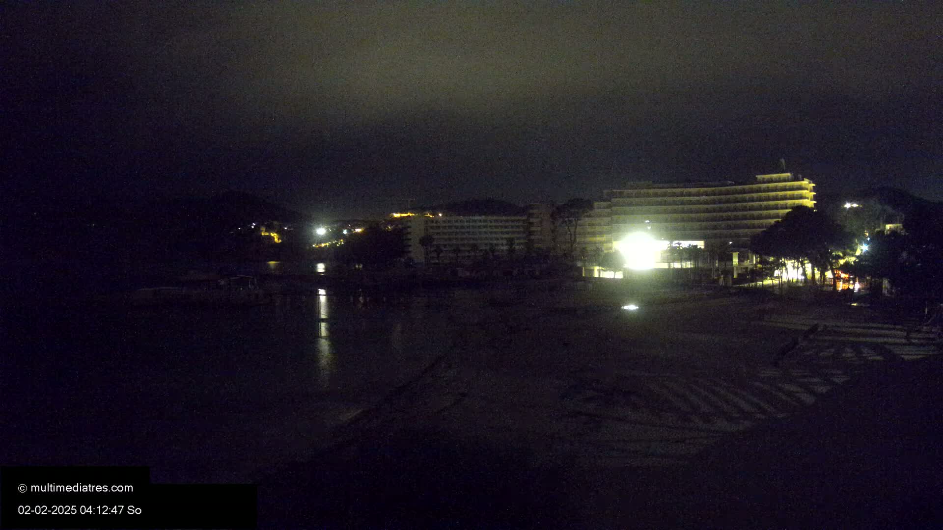 A dark, nighttime beach scene shows a brightly lit hotel and some surrounding buildings across a calm body of water.