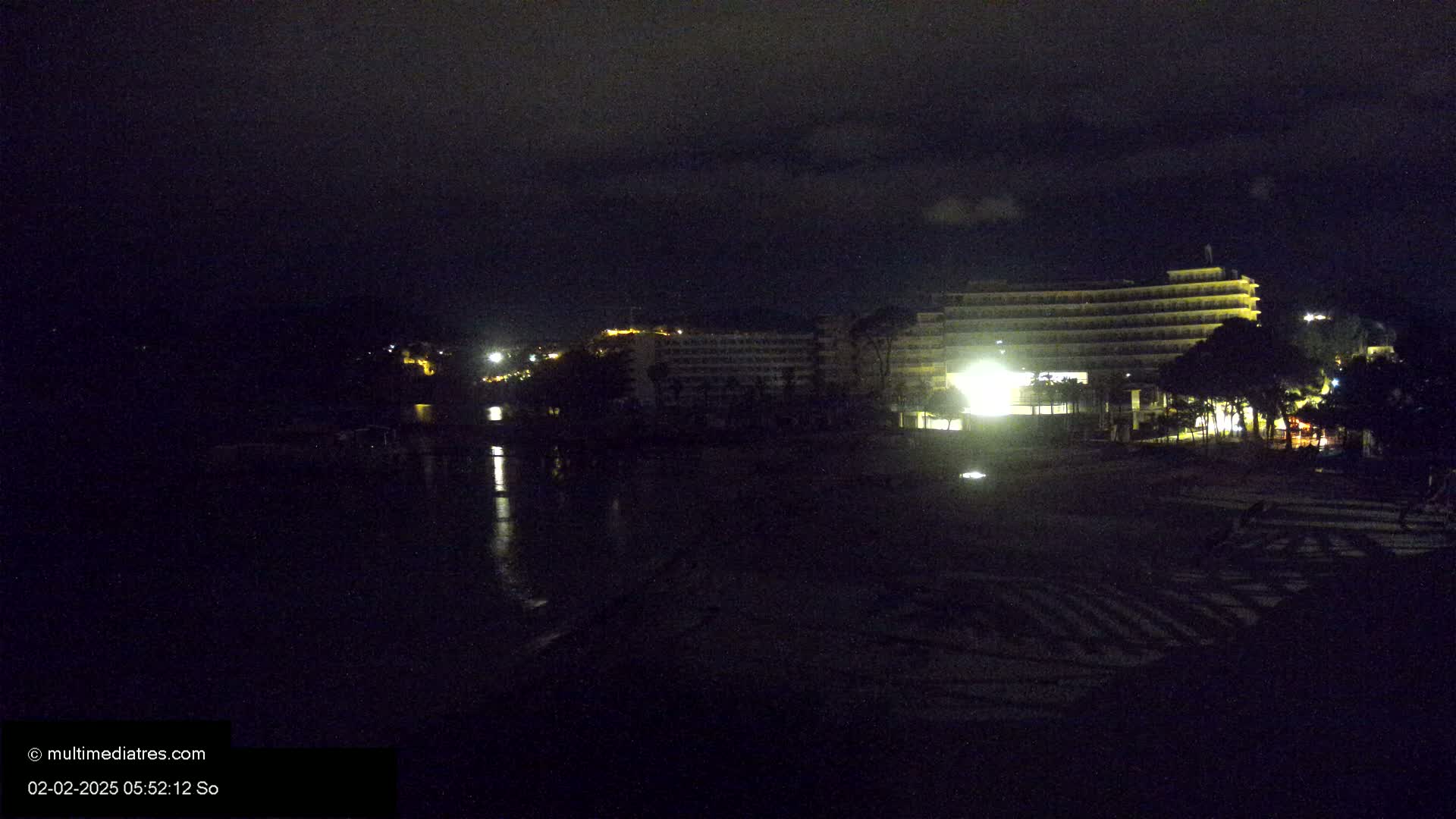 A dark, nighttime beach scene shows a brightly lit hotel in the background, and a dark, calm body of water reflecting some light in the foreground.