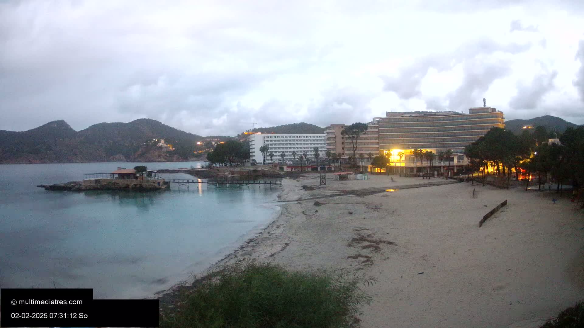 A sandy beach at dusk, with calm, light-blue water,  two hotels, and low mountains in the background under an overcast sky.