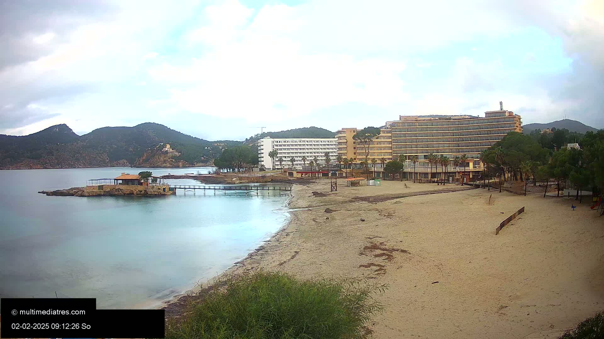 A sandy beach stretches towards two hotels, a small island with a building, and calm, light-blue water under an overcast sky.