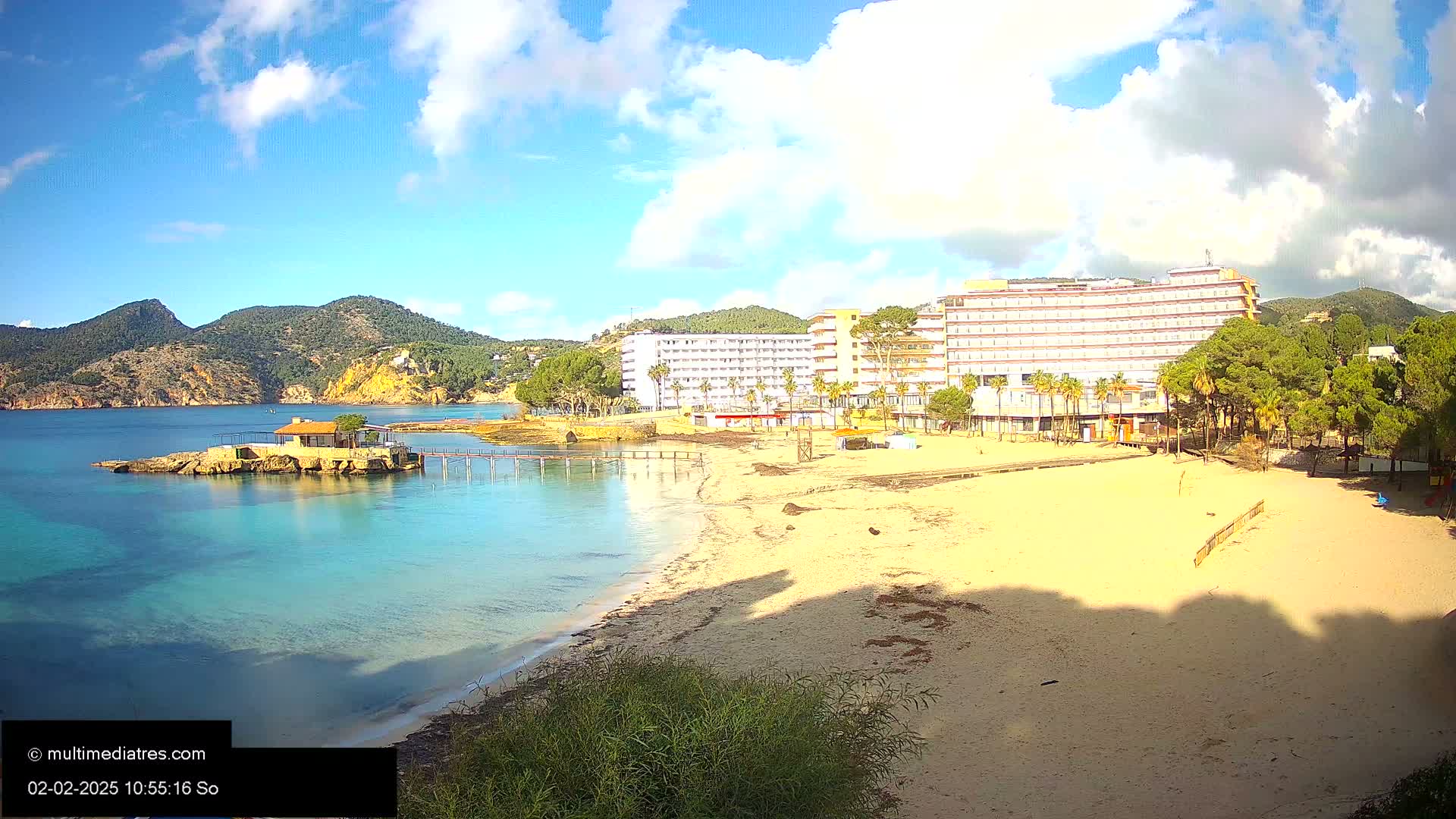 A sandy beach with turquoise water, a small building on a rocky outcrop, and a large hotel behind it under a partly cloudy sky.