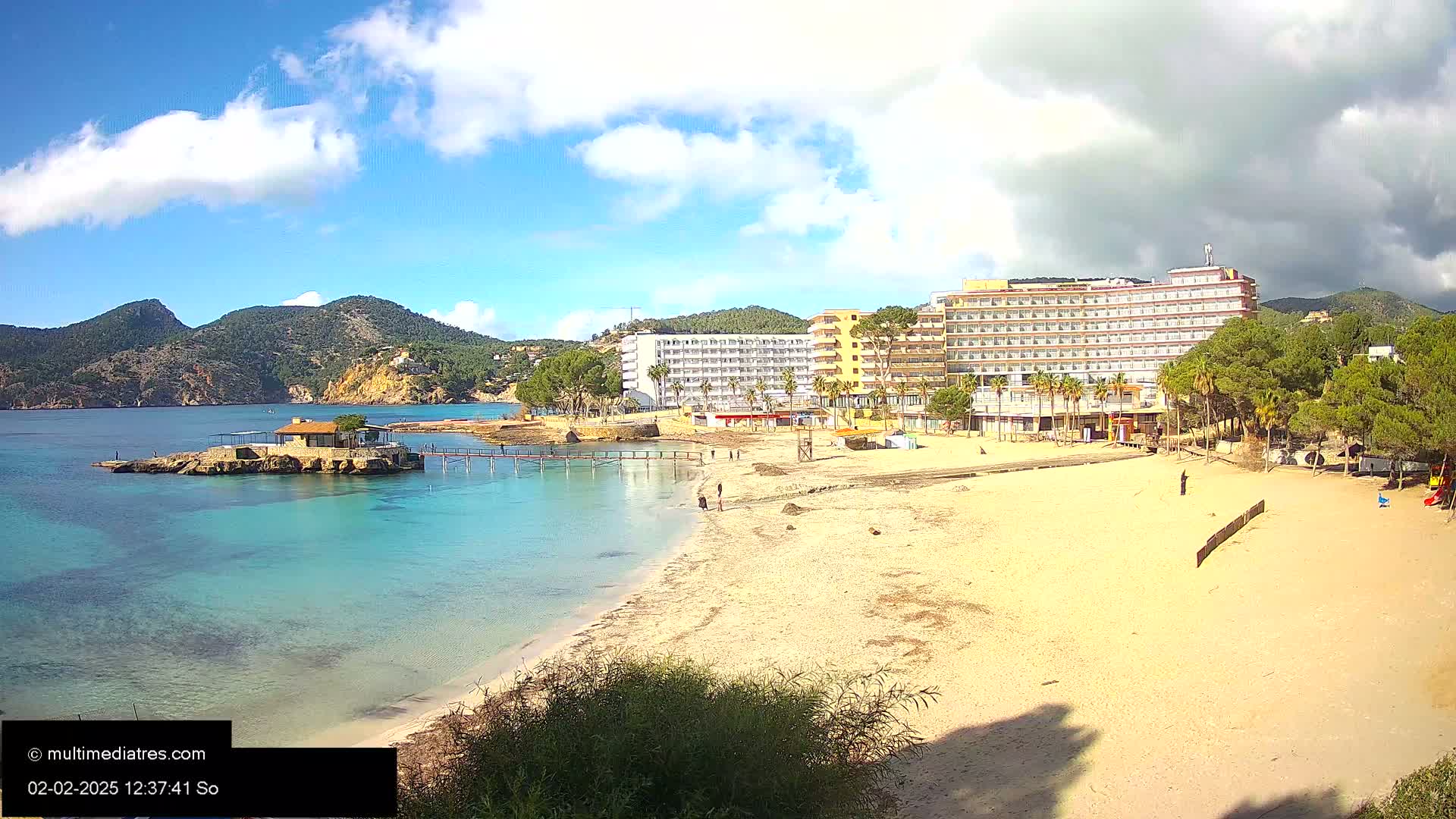 A sandy beach with turquoise water, a small island with a building, a large hotel, and some palm trees under a partly cloudy sky.