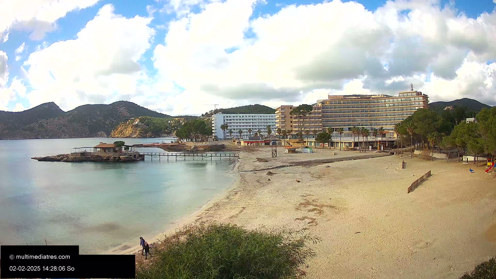 A sandy beach with a small building on a rock in the water, hotels, and mountains in the background under a partly cloudy sky.