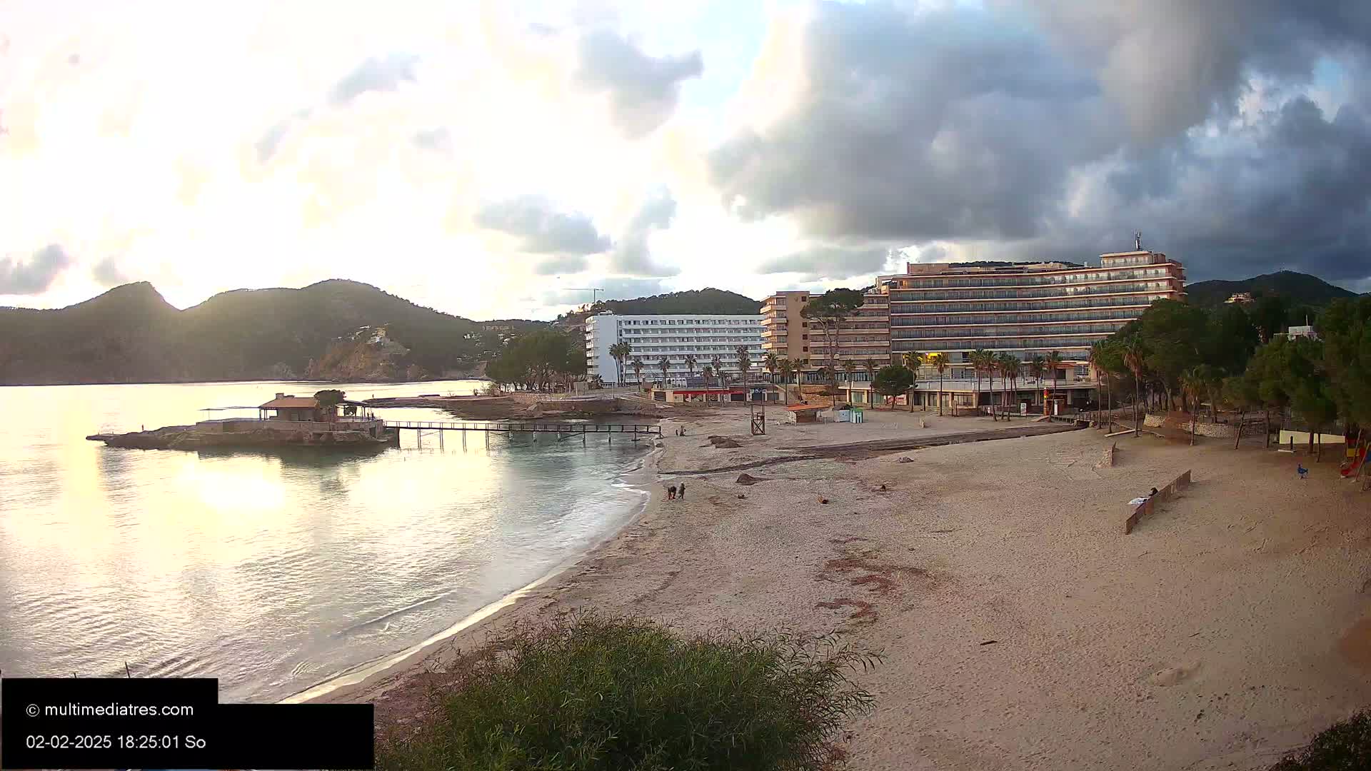 A sandy beach with a calm sea, two hotels, and hills under a partly cloudy sky.