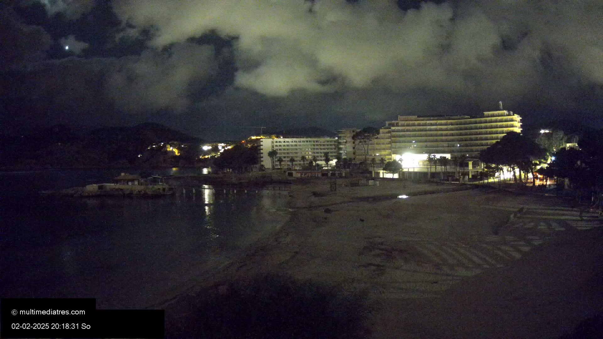 A dark, cloudy night scene shows a beach with a few illuminated hotels and buildings behind it, and calm water in front of it.