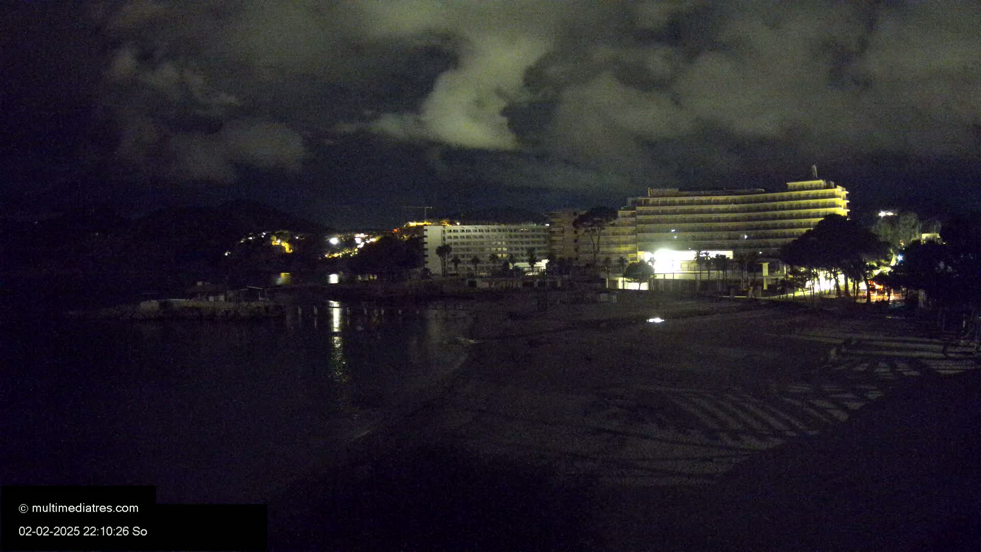 A dark, cloudy night scene shows a beach with illuminated hotels and trees, and calm water reflecting some lights.