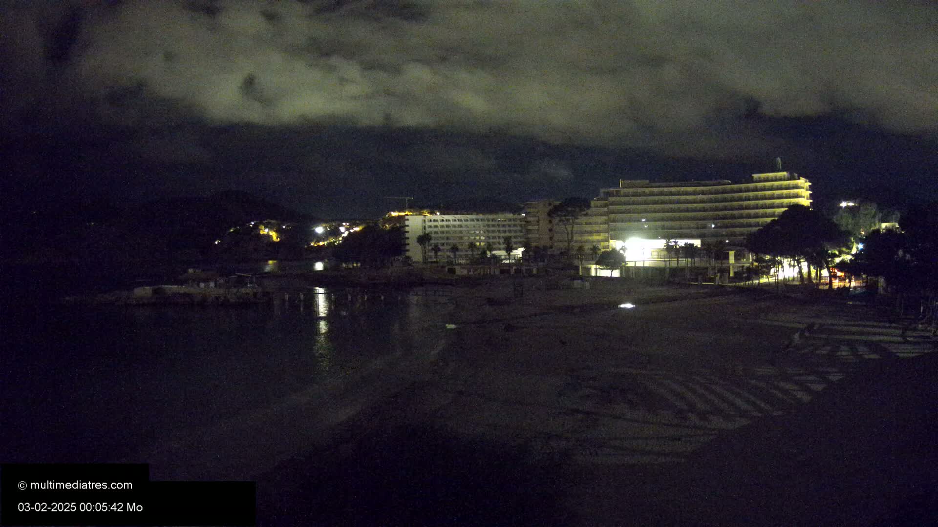 A nighttime view of a beach with a large hotel illuminated against a dark, cloudy sky.