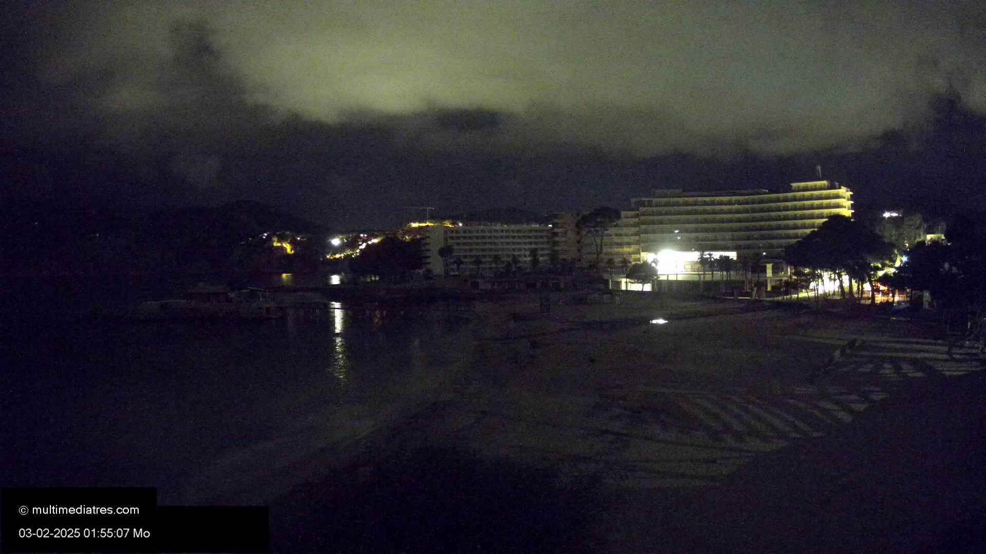 A dark, nighttime beach scene shows a large, illuminated hotel building and calm water under a cloudy sky.