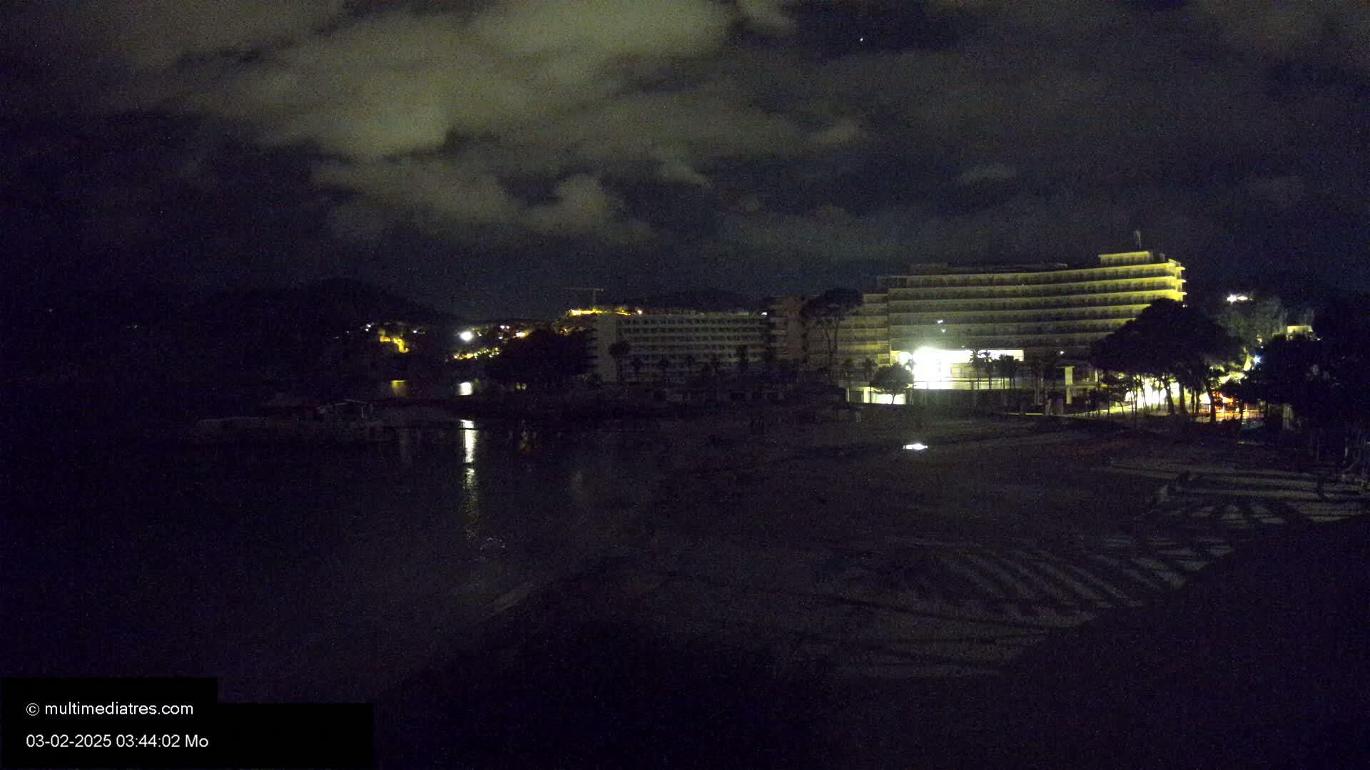 A dark, cloudy night scene shows a beach with a large, illuminated hotel in the background, and calm water reflecting some light.
