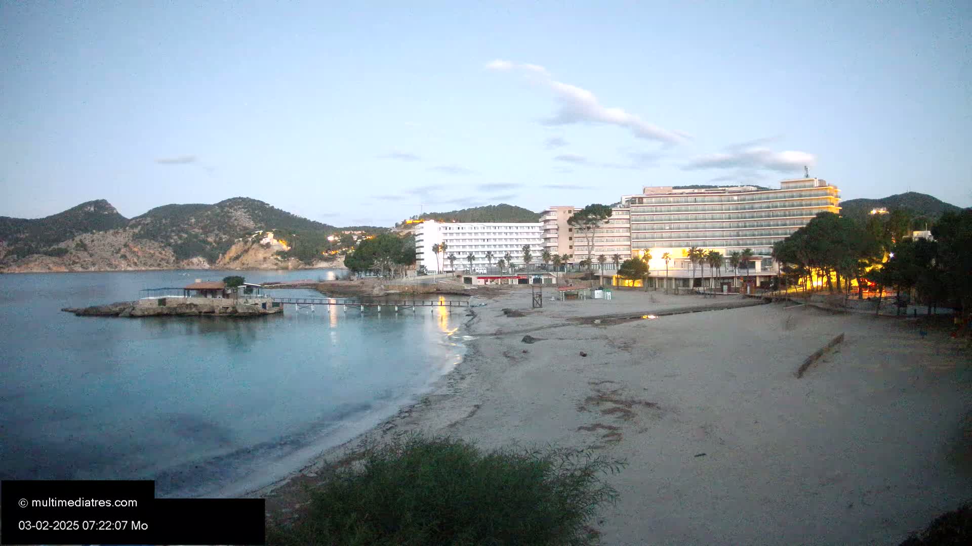 A sandy beach at twilight is situated near two large hotels, with calm water and a small island with a structure visible in the bay, backed by dark hills.