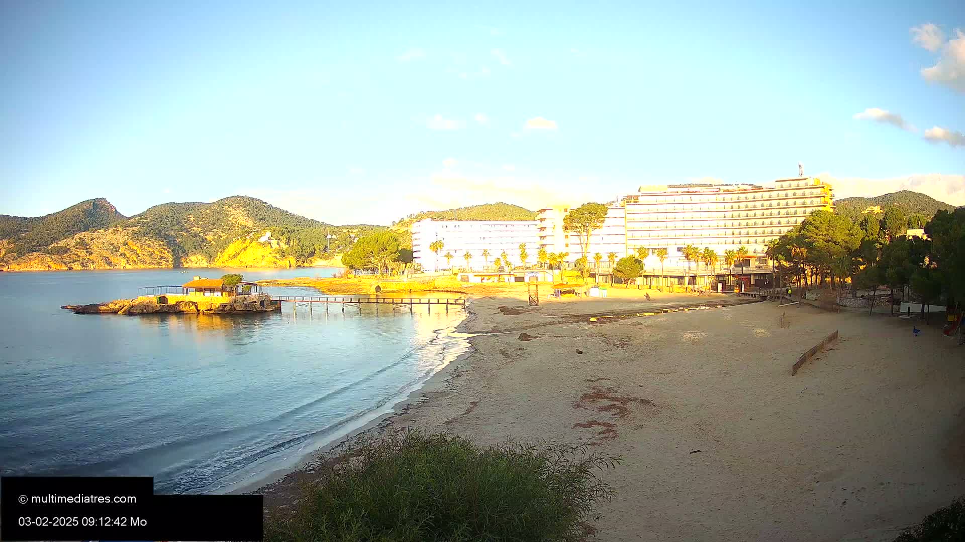 A sandy beach with calm, clear water and a large hotel in the background under a mostly clear sky.
