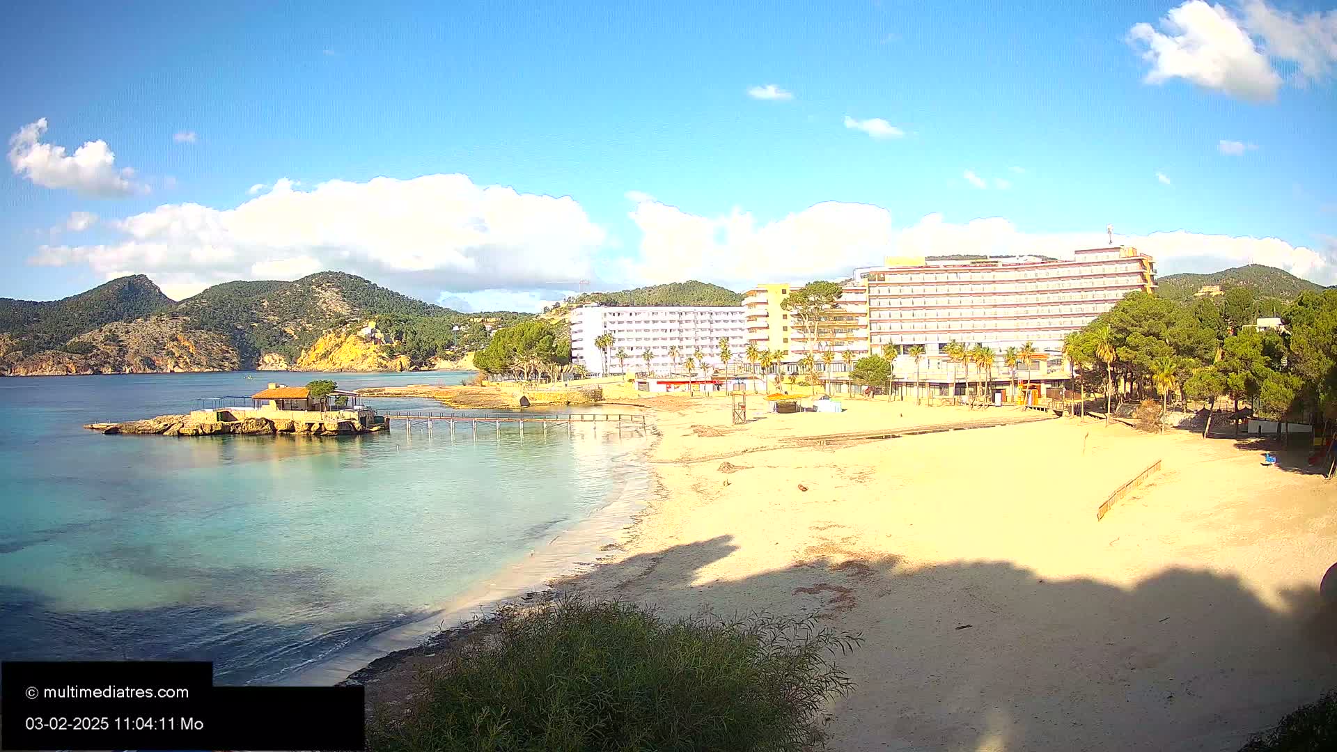 A sandy beach stretches towards a large hotel, with a small island and calm, clear water in the foreground under a partly cloudy sky.