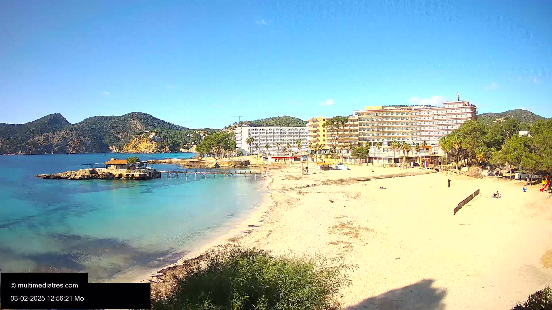 A sandy beach with a small building on a rocky outcrop in clear, shallow turquoise water, next to a large hotel complex under a bright, sunny sky.