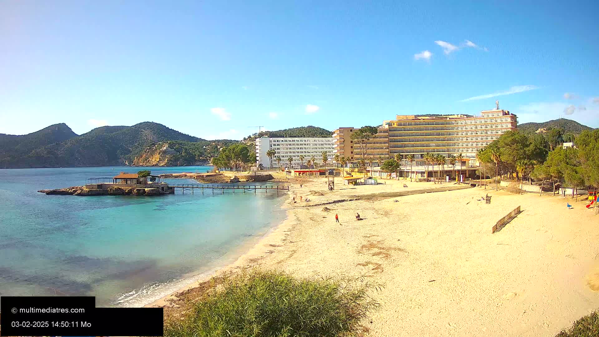 A sandy beach with turquoise water, a small building on a rock jetty, a large hotel, and some palm trees under a clear blue sky.