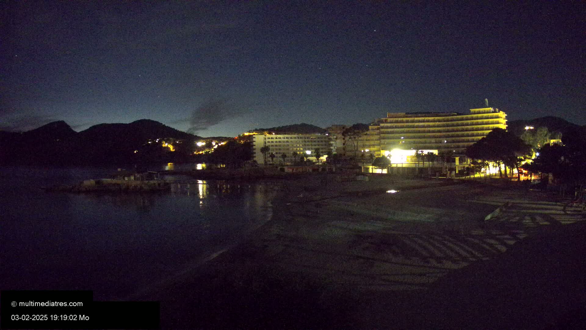 A dark, nighttime beach scene shows a brightly lit hotel complex against a backdrop of silhouetted hills under a starry sky.