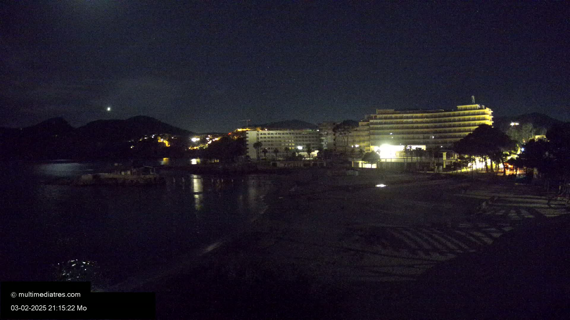 A moonlit nighttime view of a beach with several illuminated hotels and trees in the background.
