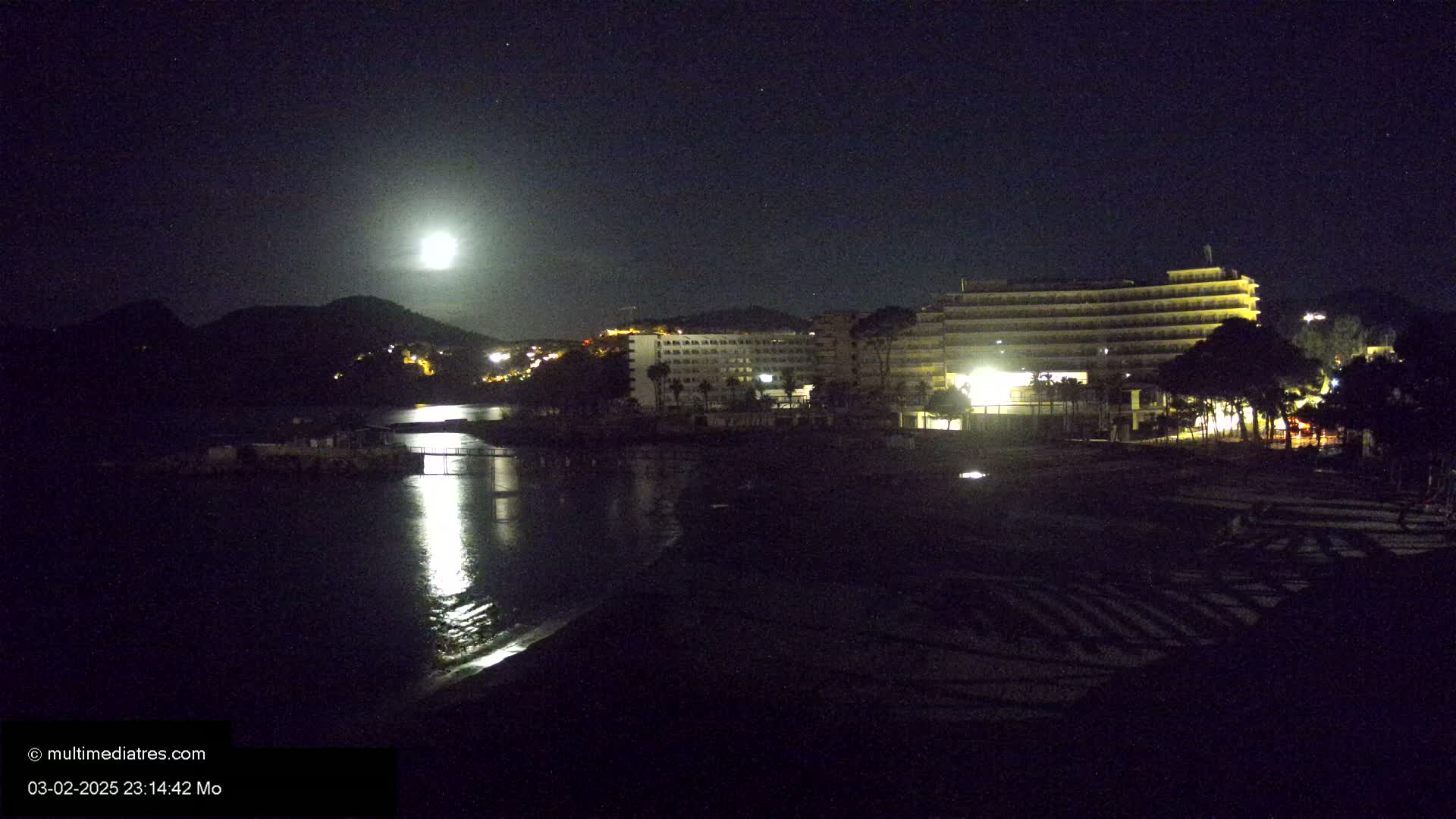 A nighttime view of a beach with a large hotel and hills in the background, illuminated by a bright moon.
