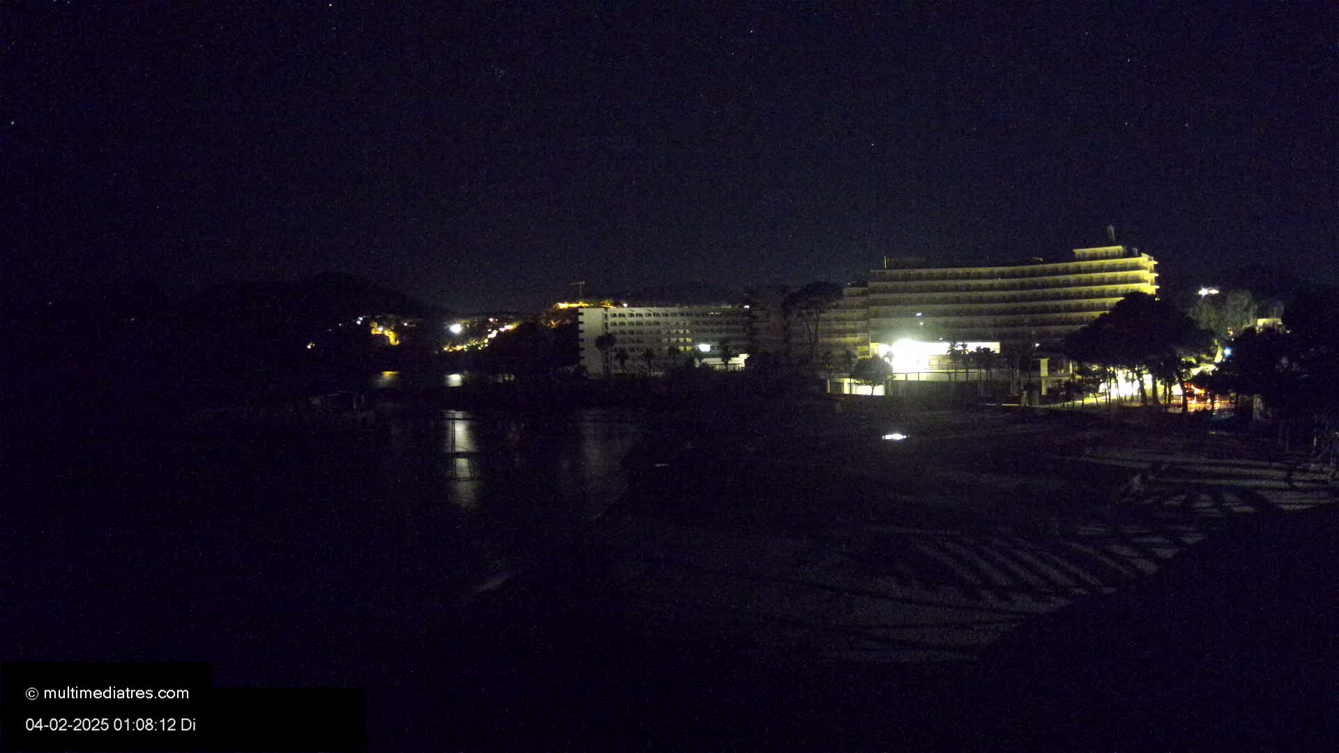 At night, under a clear, starry sky, several illuminated hotel buildings are situated near a dark body of water, with some vegetation in the foreground.