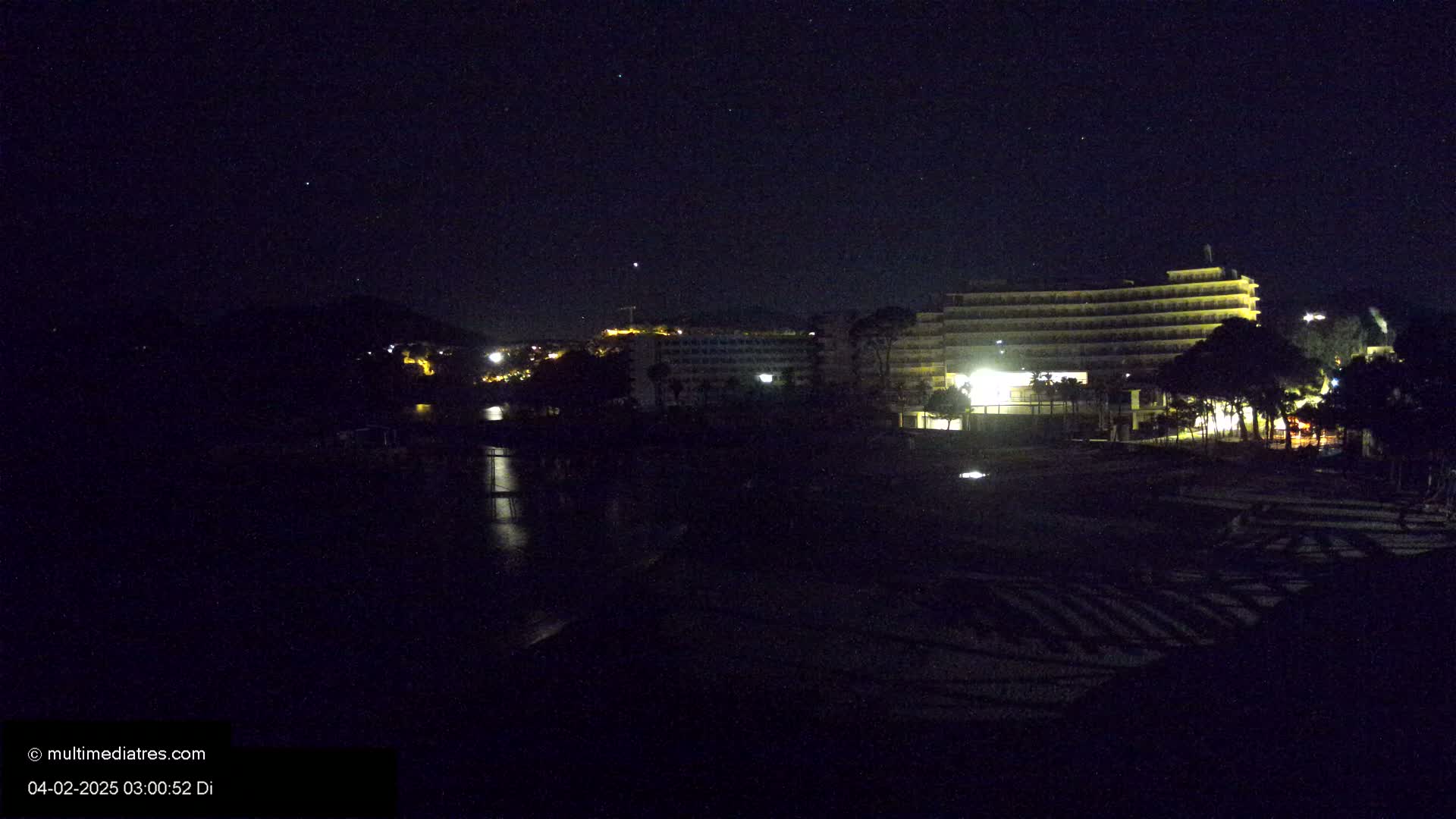 A nighttime view of a brightly lit hotel complex situated near a dark body of water under a clear, starlit sky.