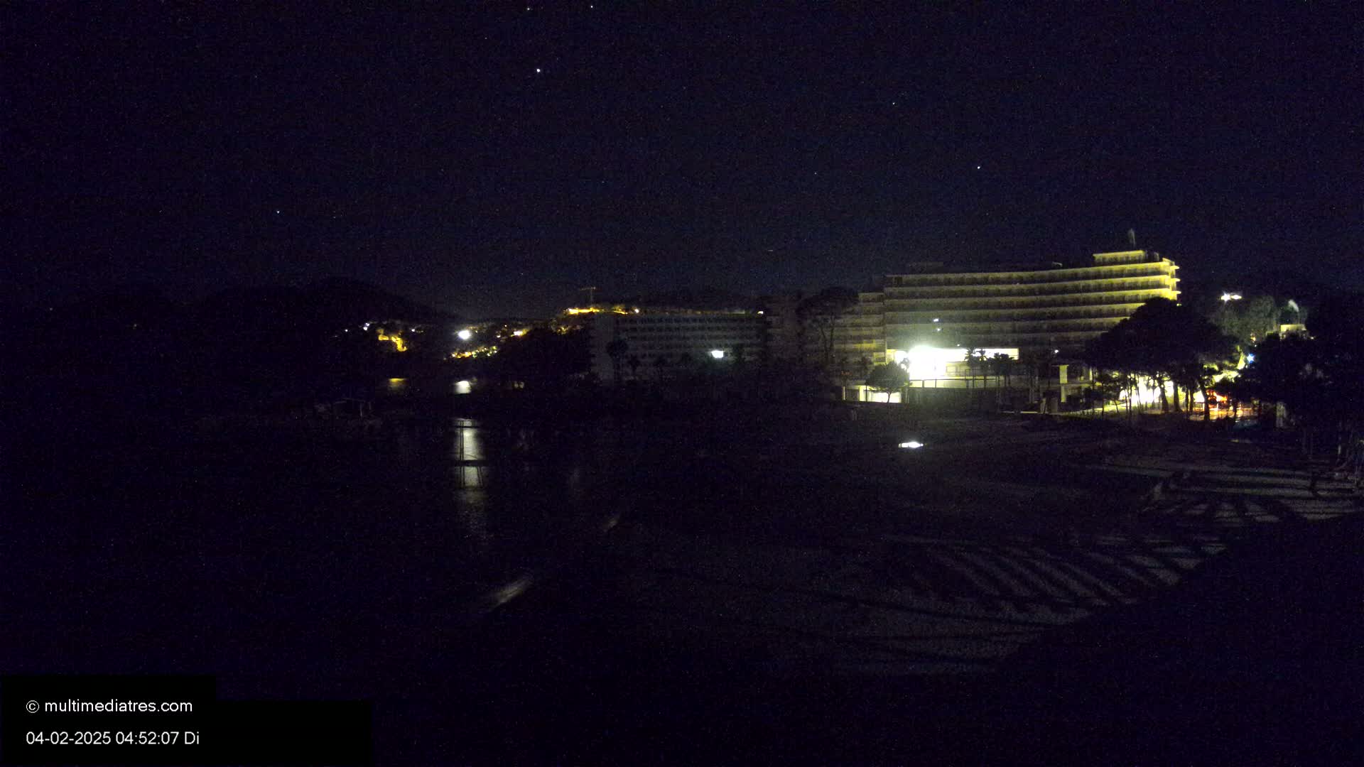 A nighttime view shows a dark beach with a brightly lit hotel complex and a calm body of water reflecting the lights.