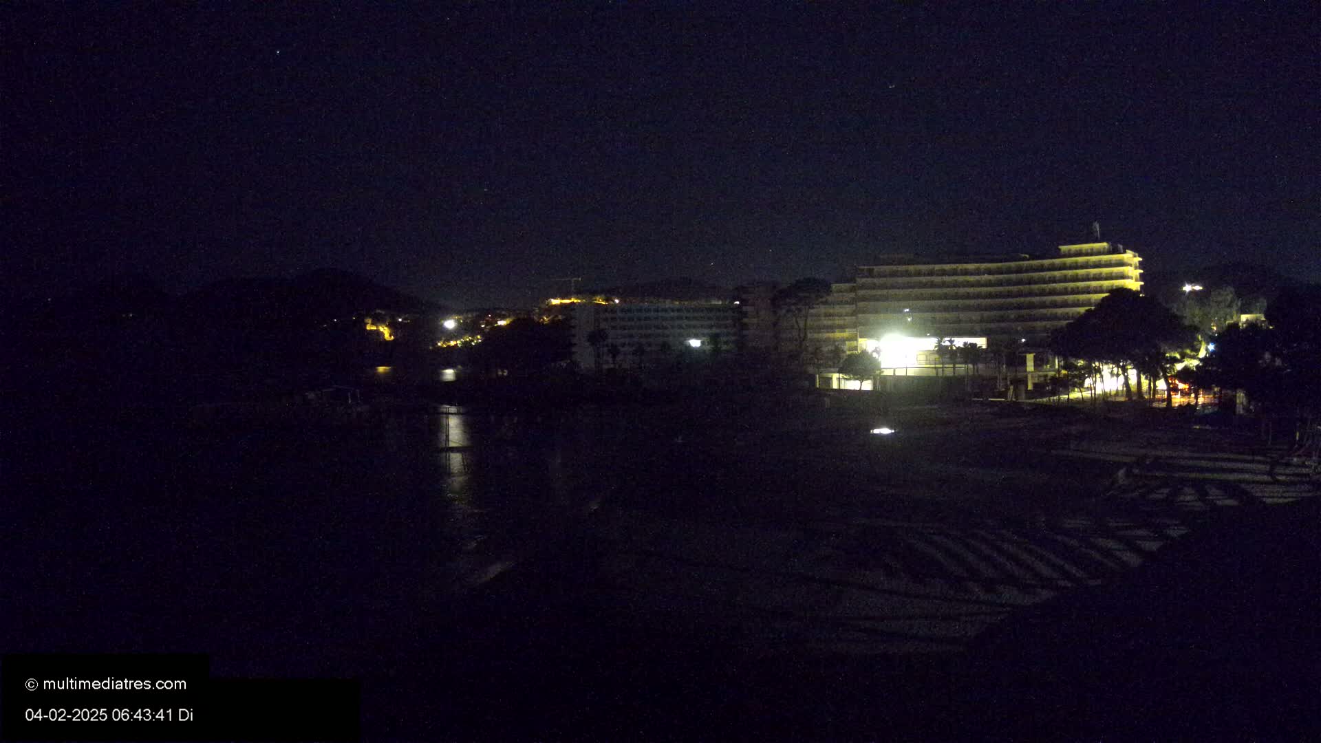 A dark, nighttime view of a beach with a multi-story hotel and trees illuminated by lights.