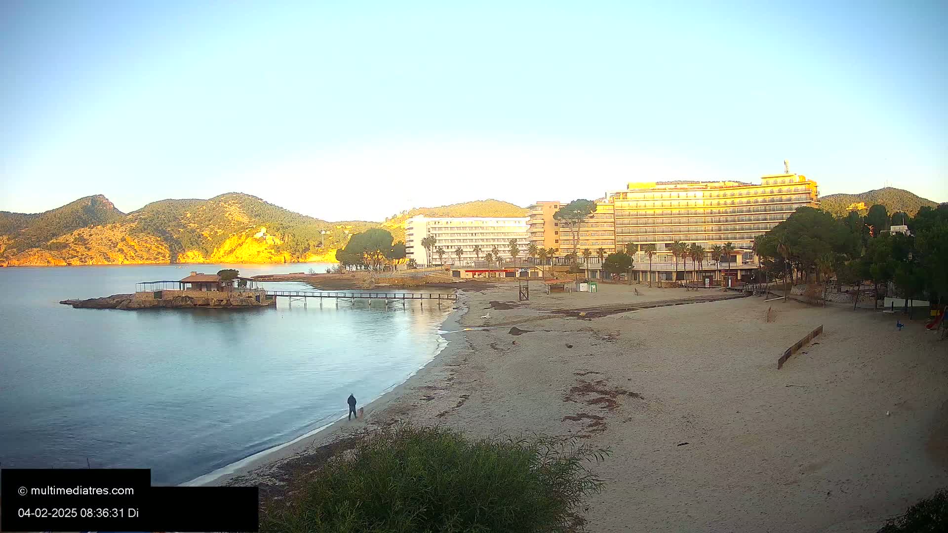 A sandy beach with calm water, a small island with a building, hotels, and hills in the background under a clear, sunny sky.