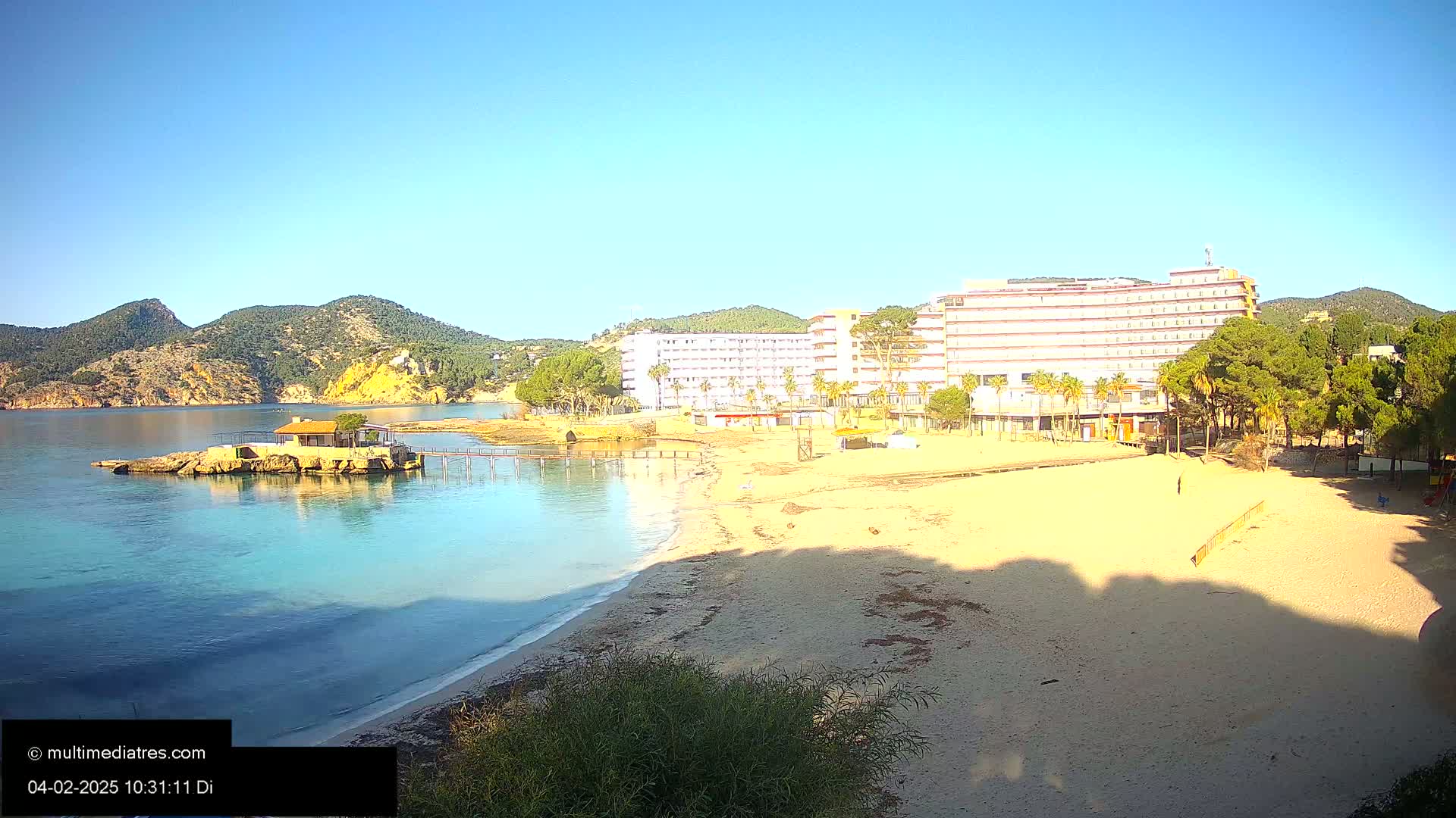 A sandy beach with calm, clear turquoise water, a small building on an island, and a large hotel behind it, all under a clear blue sky.