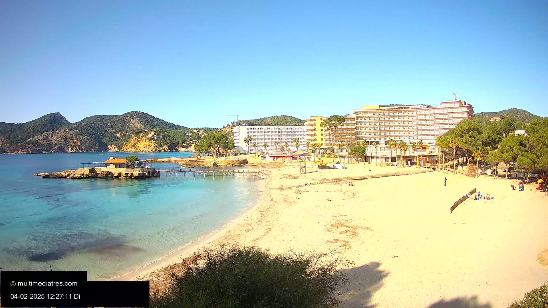 A sandy beach with clear turquoise water, a small island with a building, and large hotels behind it under a sunny sky.