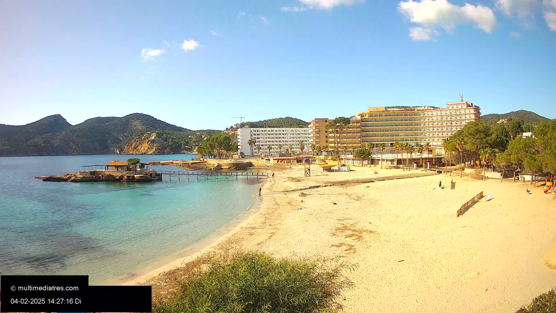 A sandy beach with a few people and a large hotel is seen next to clear turquoise water, backed by green hills under a mostly sunny sky.