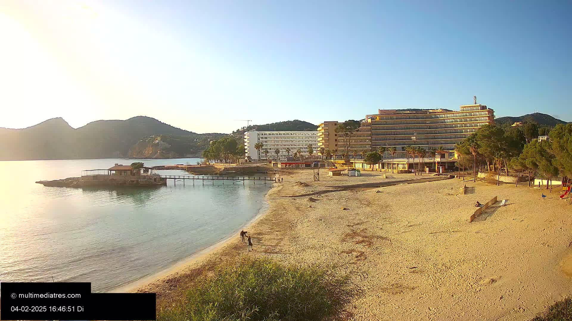 A sandy beach with several hotel buildings in the background stretches along a calm body of water under a clear, sunny sky near some low mountains.