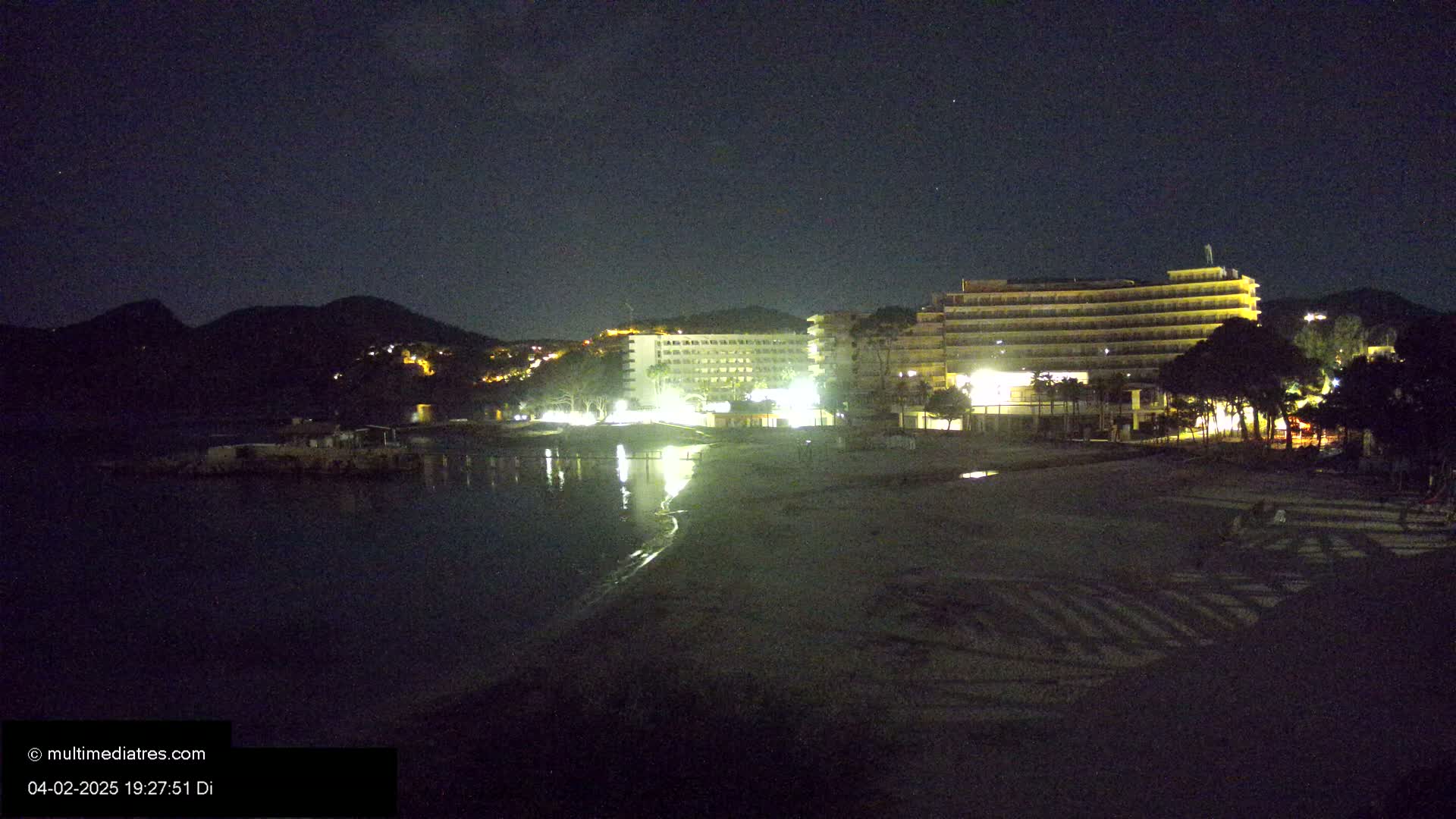 A nighttime view of a beach with illuminated hotels and dark mountains in the background under a clear, starry sky.