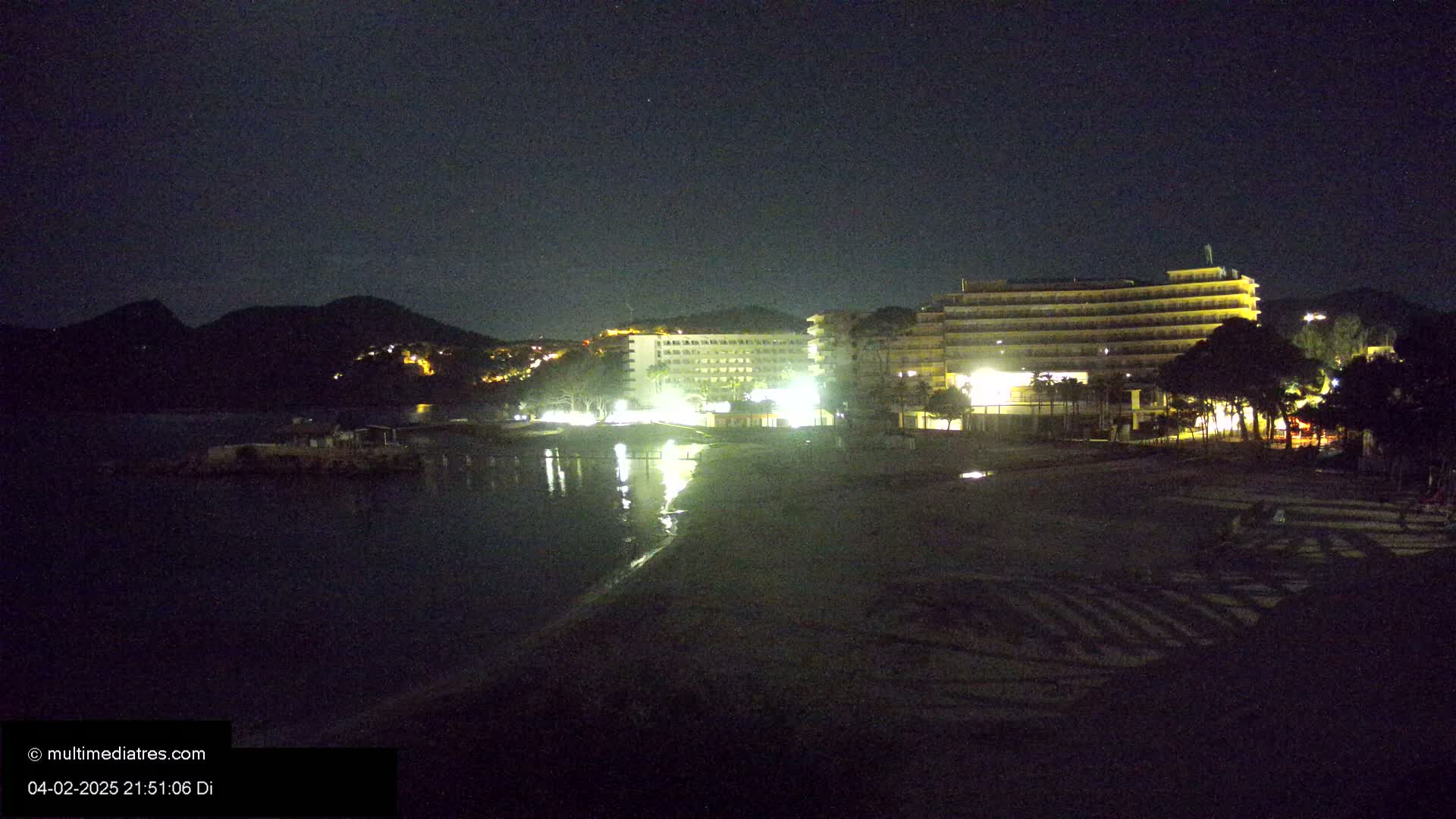 At night, illuminated hotels are situated on a beach next to a dark body of water and a hill.