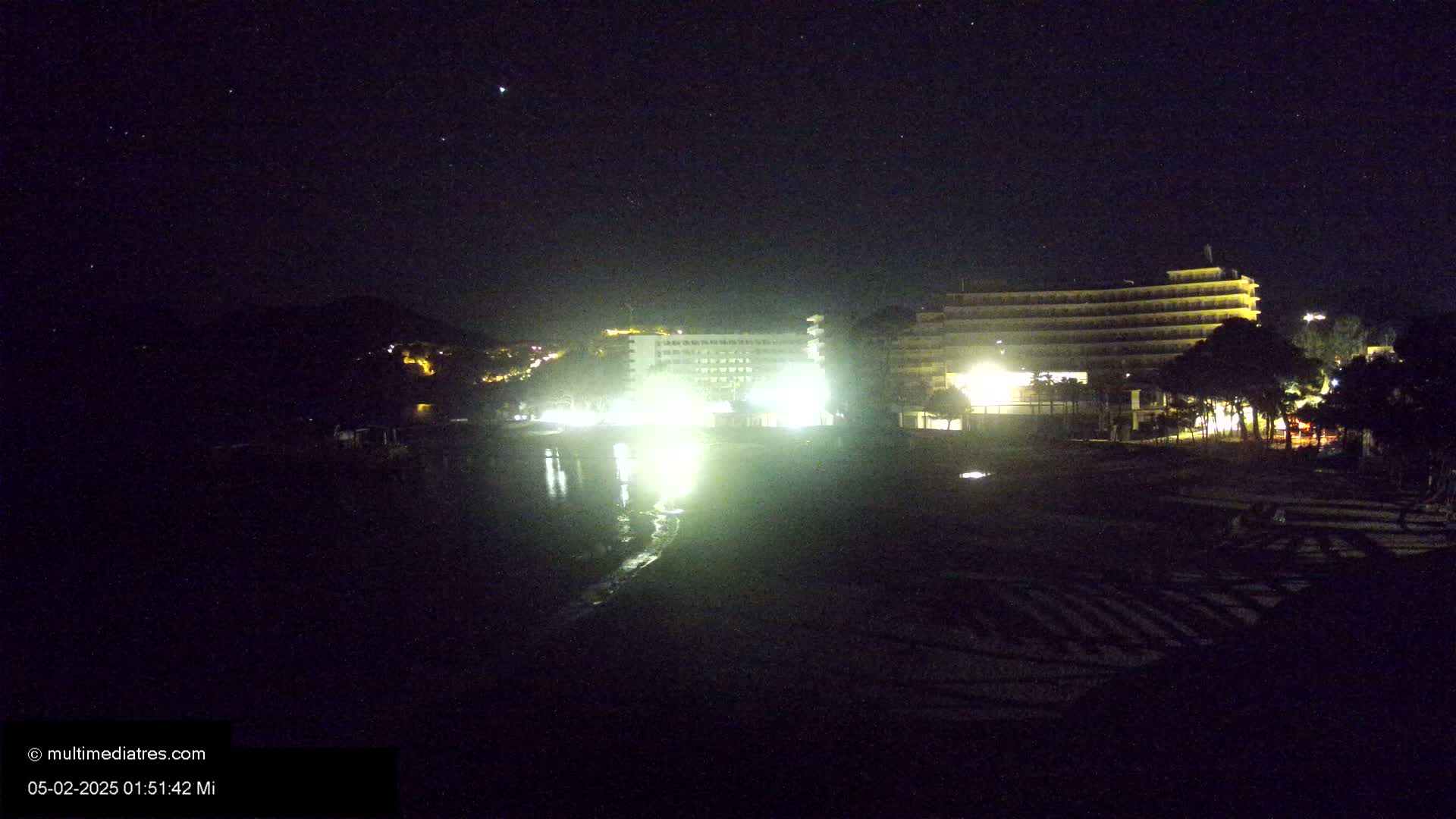 Two brightly lit hotels are situated on a dark, nighttime beach under a clear, starry sky.