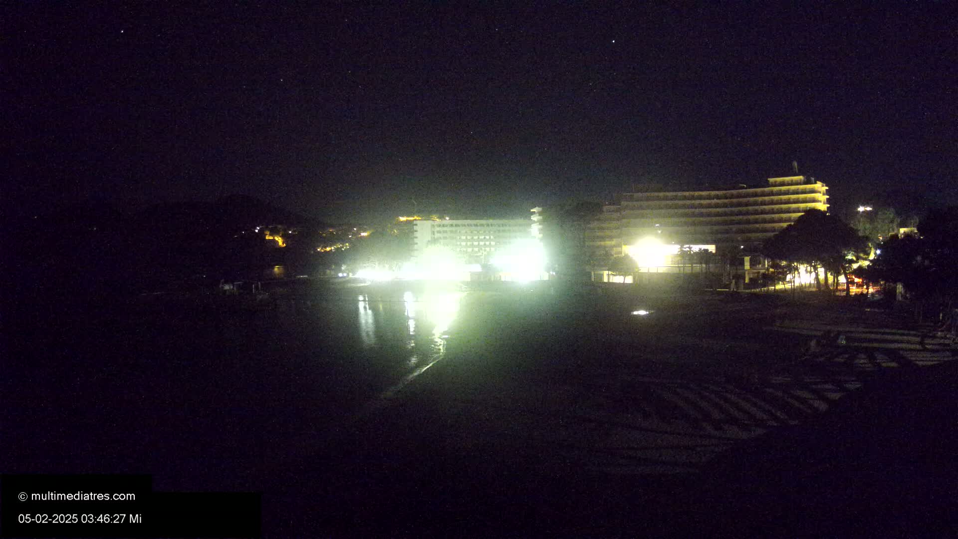 At night, two brightly lit hotels stand near a dark beach and calm body of water under a clear, starry sky.
