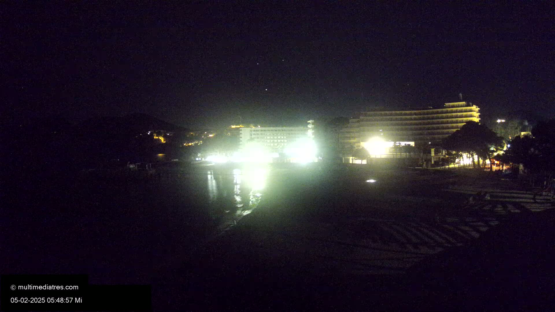At night, brightly lit hotels are situated near a dark beach and calm body of water under a clear, starry sky.