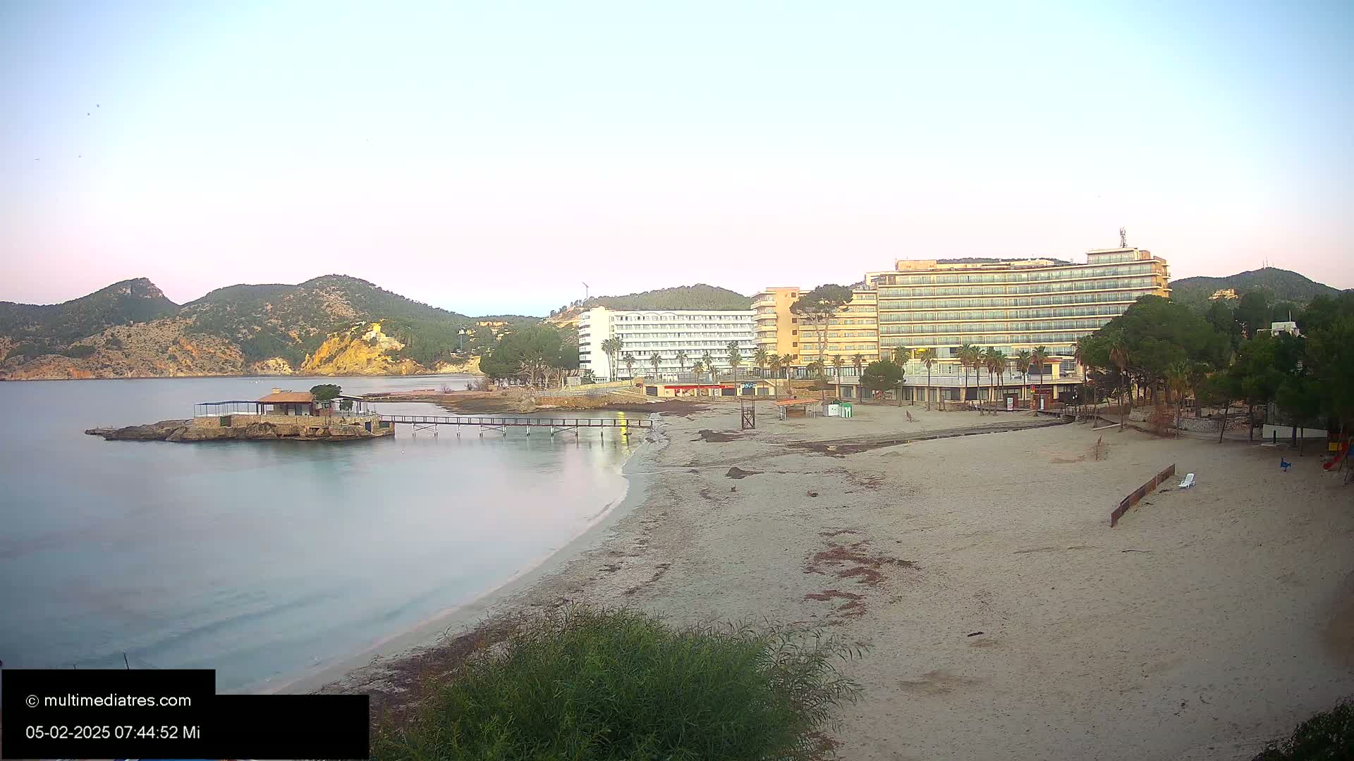 A sandy beach with calm, clear water, a small island with a building, and several hotels under a pale dawn sky.
