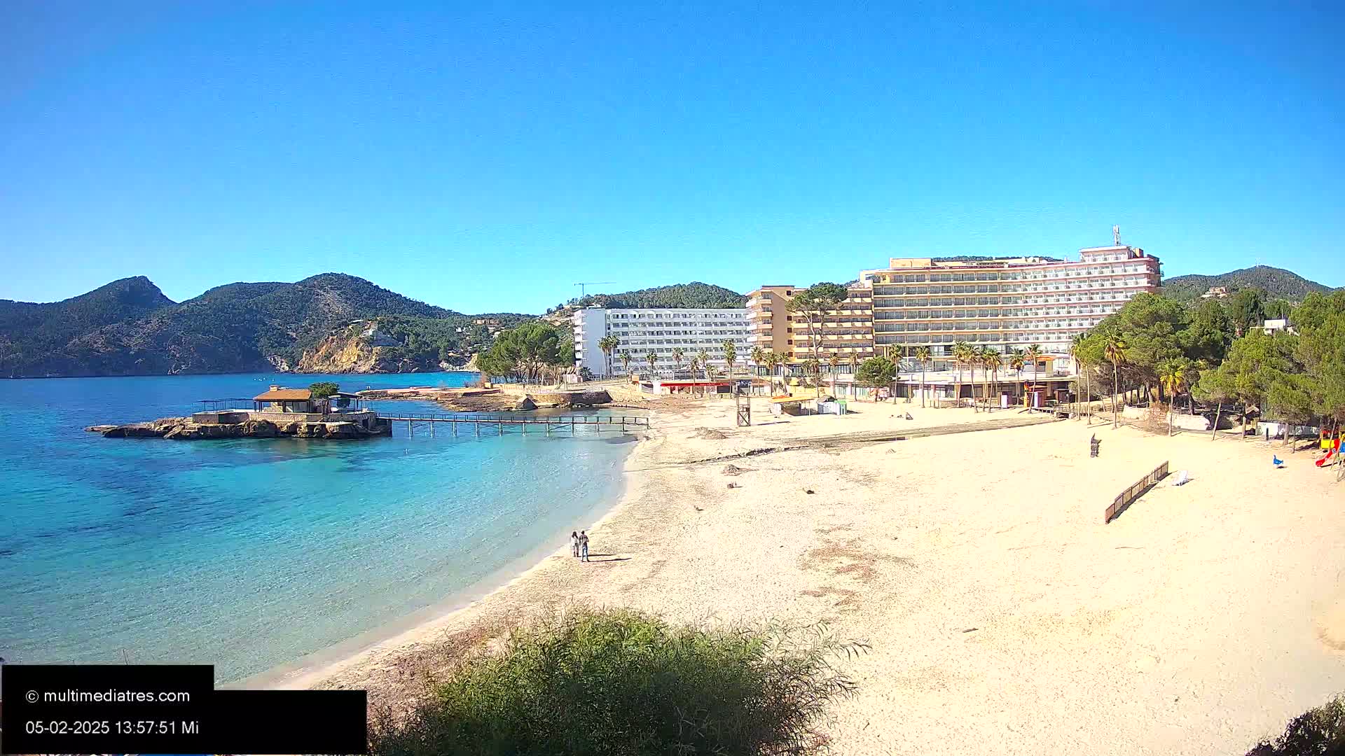 A sandy beach with a small building on a rock in the water, a hotel, and pine trees, all under a clear blue sky.