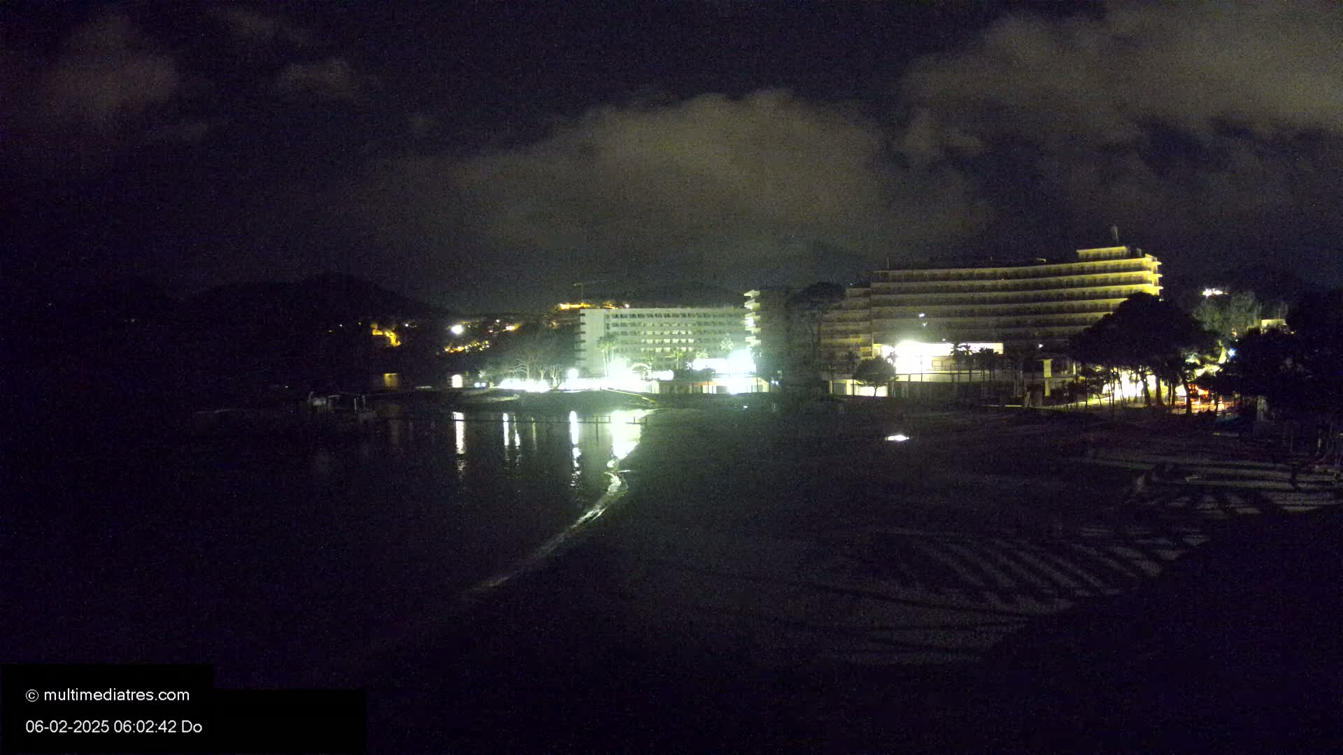 Two brightly lit hotels are situated on a dark beach at night under a cloudy sky.