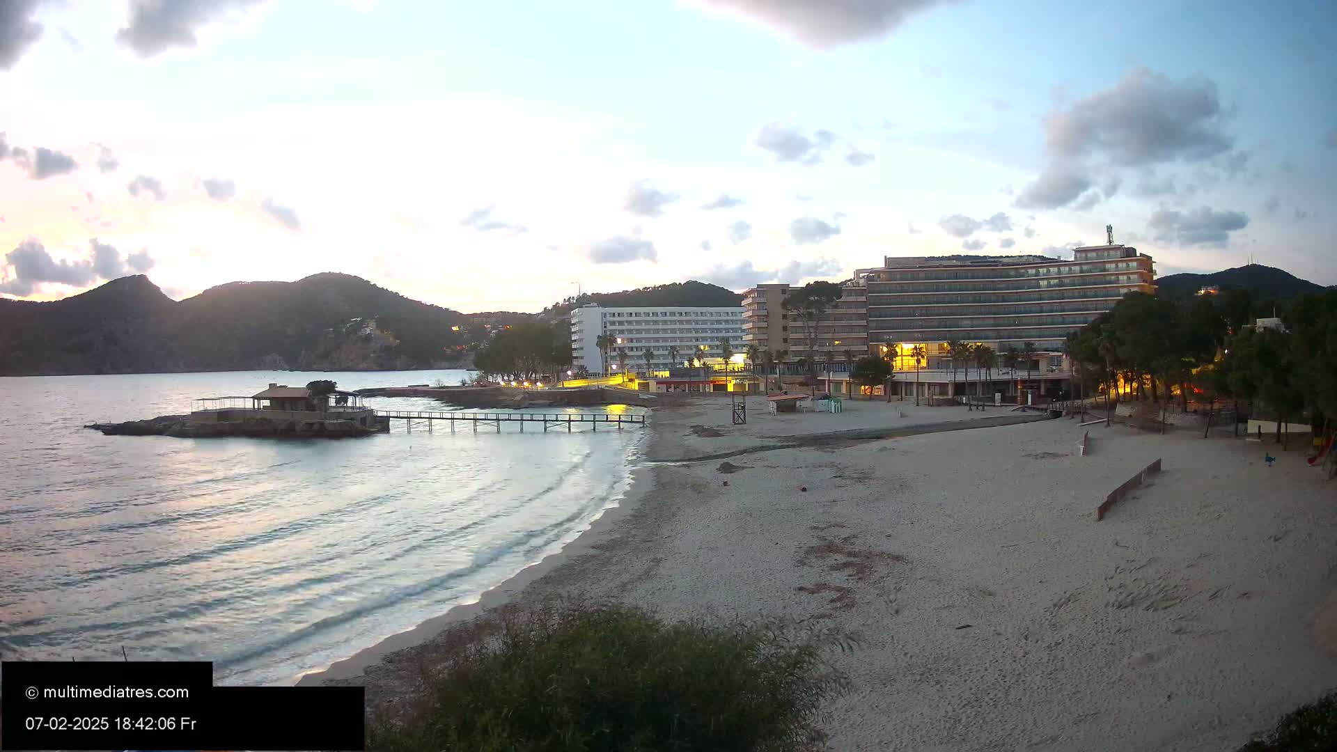 A sandy beach at dusk, with calm water lapping the shore, shows a pier extending from a small building on a rock, and several hotels and buildings in the background under a cloudy sky.