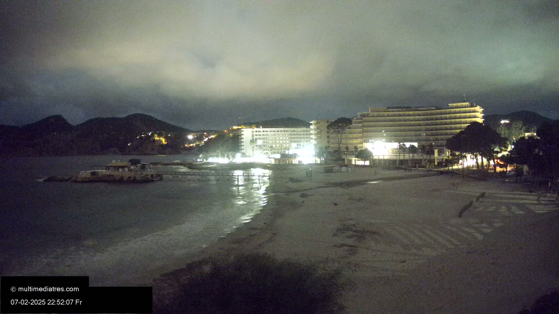 A nighttime beach scene shows a sandy shore with lit hotels and hills in the background under a cloudy sky.