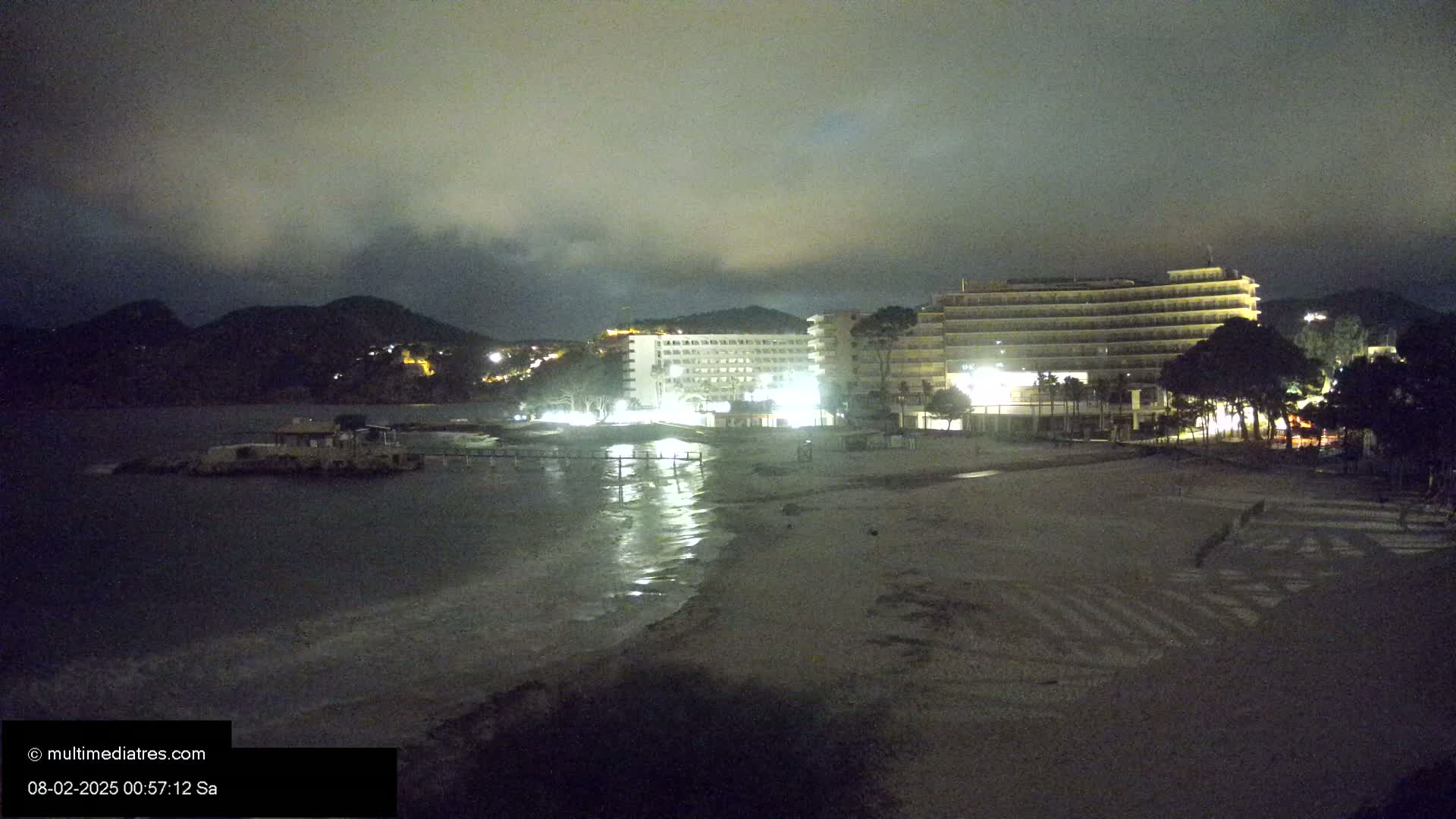 A beach at night under a cloudy sky shows a lit-up hotel and other buildings across the sand from the water.
