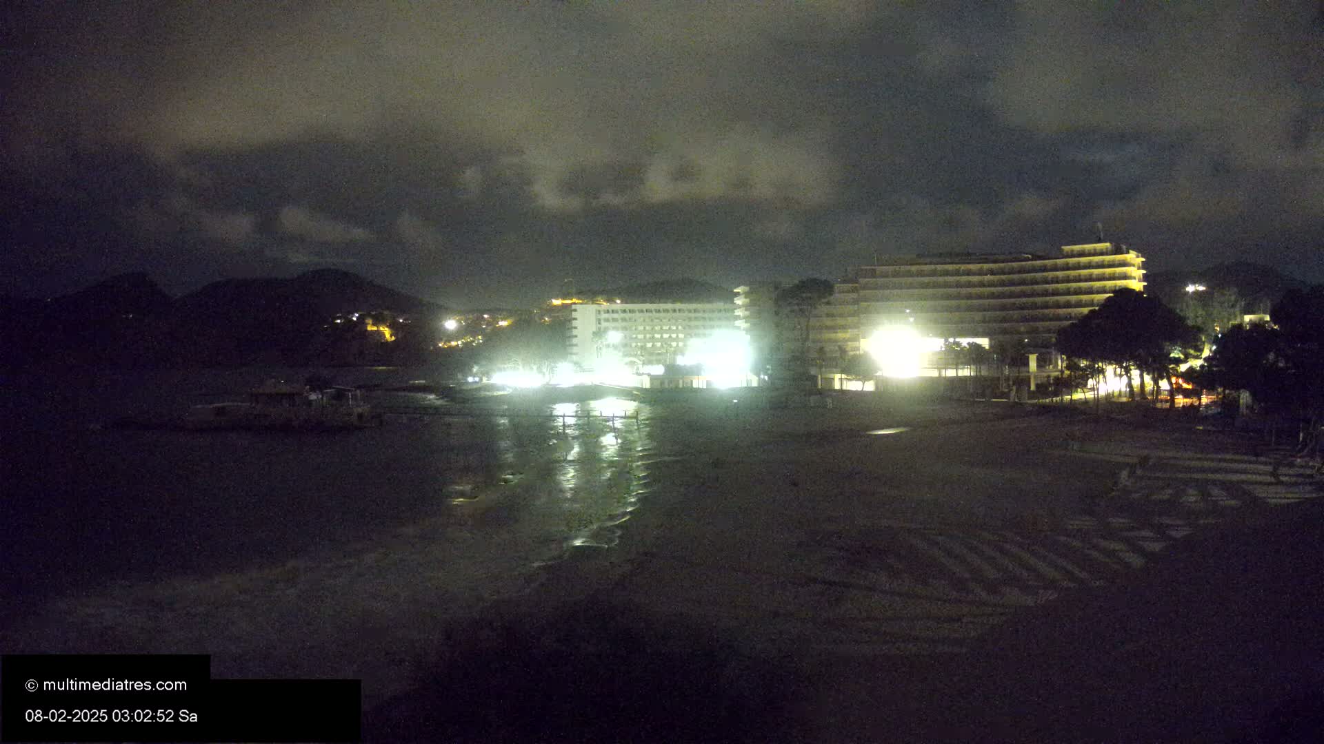 At night, under a cloudy sky, illuminated hotels are situated near a dark beach with gentle waves lapping the shore.