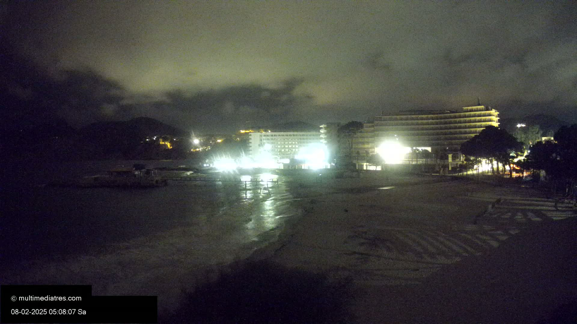 At night, under a cloudy sky, two brightly lit hotels are situated near a dark beach with a wet shoreline.