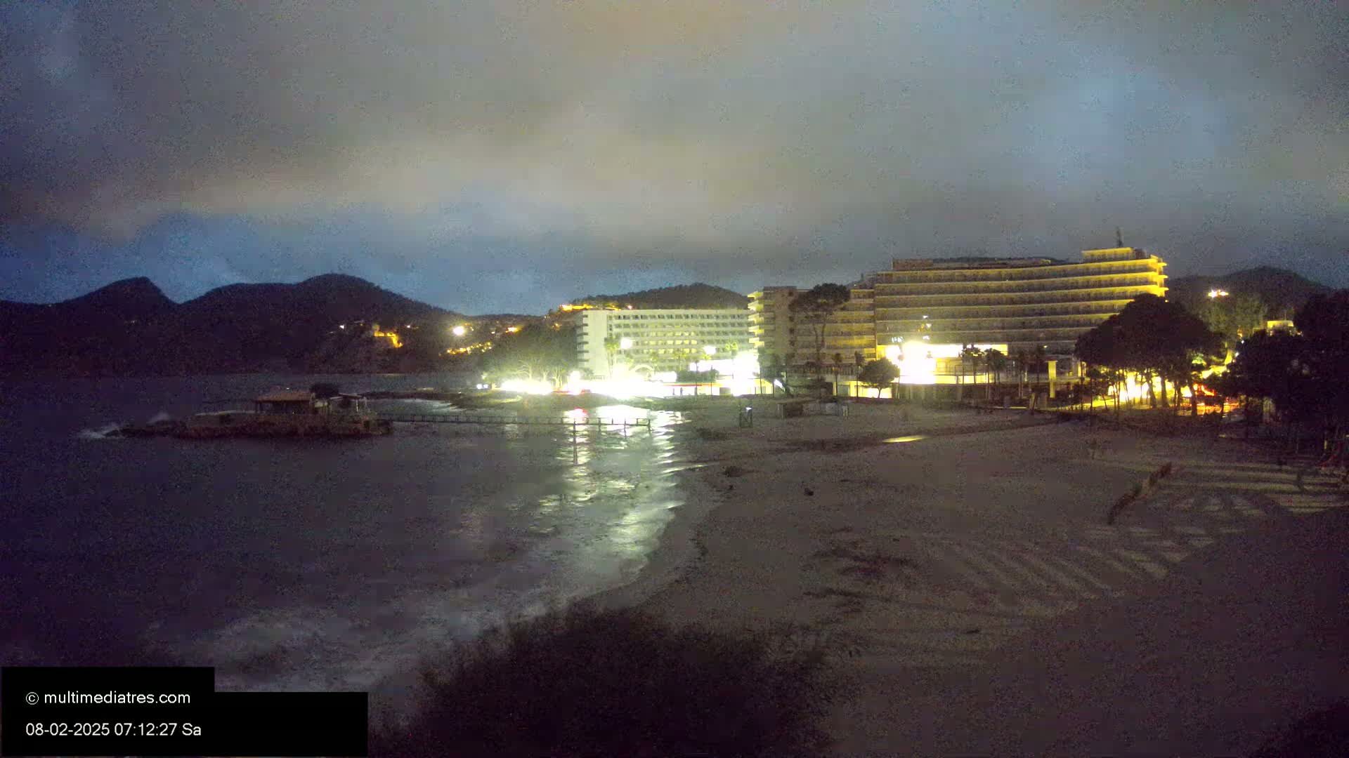 A nighttime view of a beach with illuminated hotels and mountains in the background under a cloudy sky.
