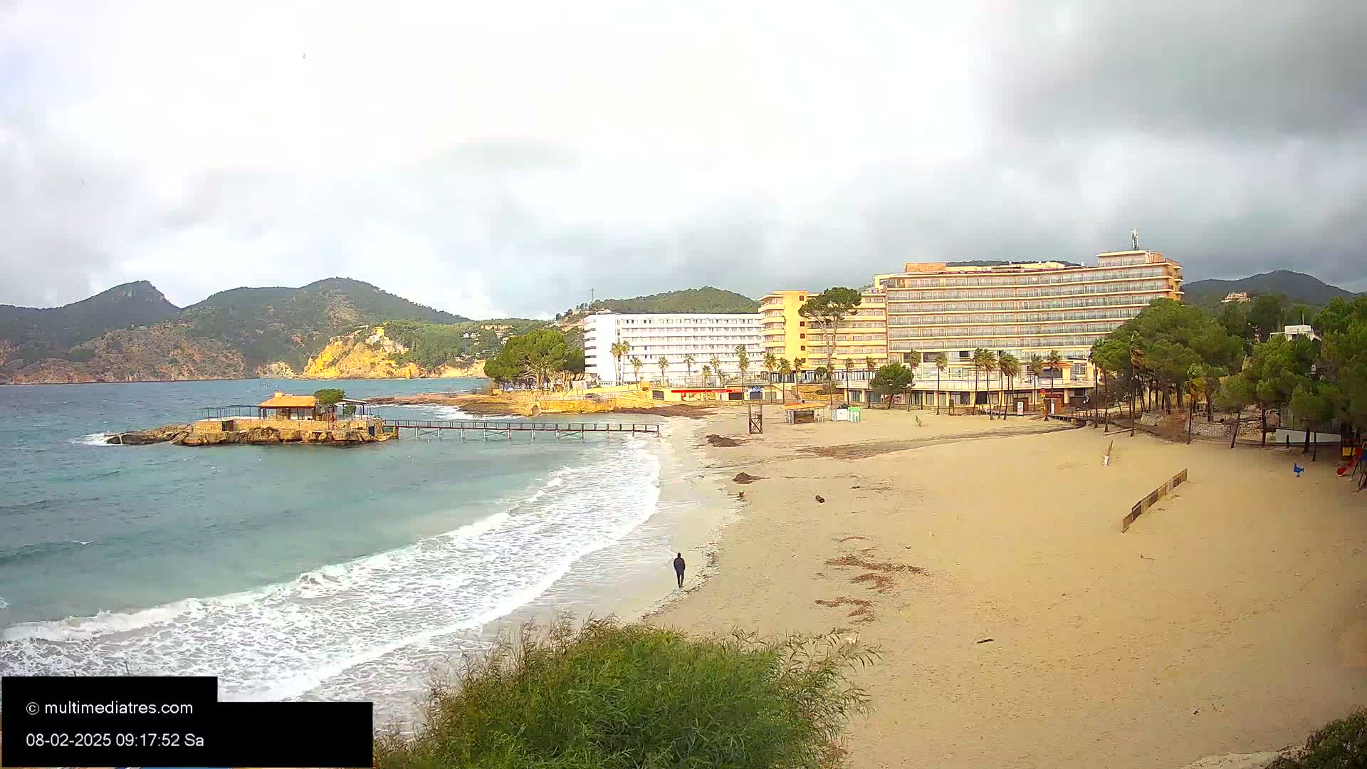 A sandy beach with a person walking on it, stretches out in front of several hotels and a small island with a building under an overcast sky.