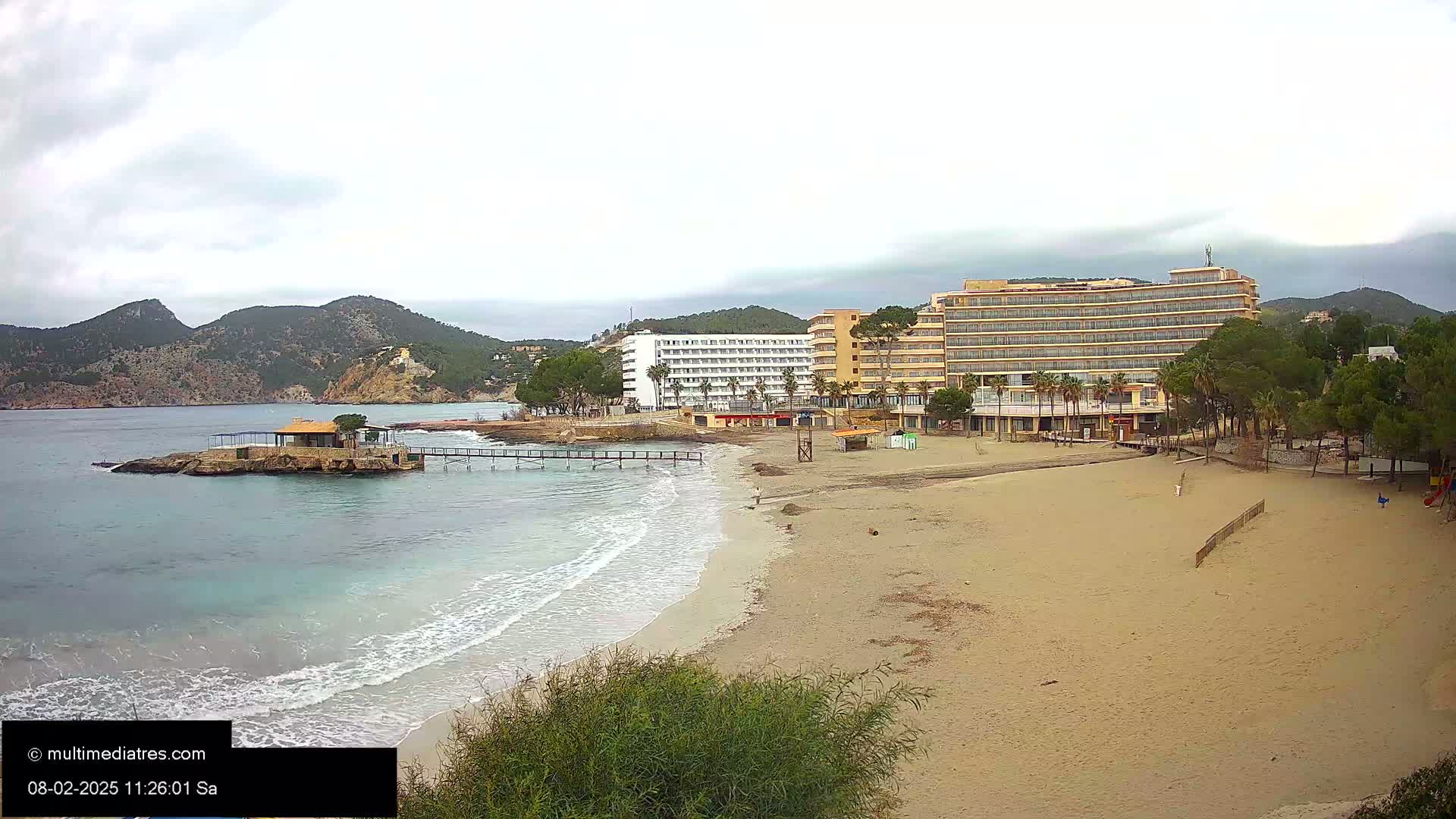 A sandy beach with gentle waves laps the shore, near two large hotels and a small island with a building, all under an overcast sky.