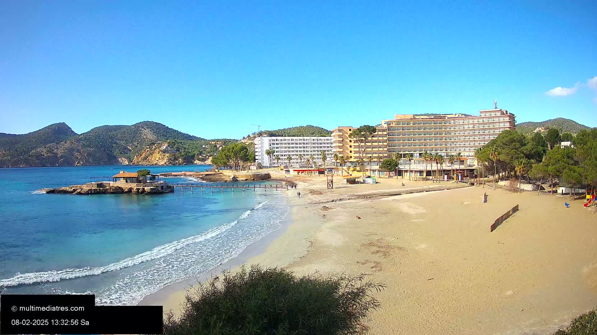 A sandy beach with light waves laps a shore near a large hotel complex, backed by green hills under a clear blue sky.